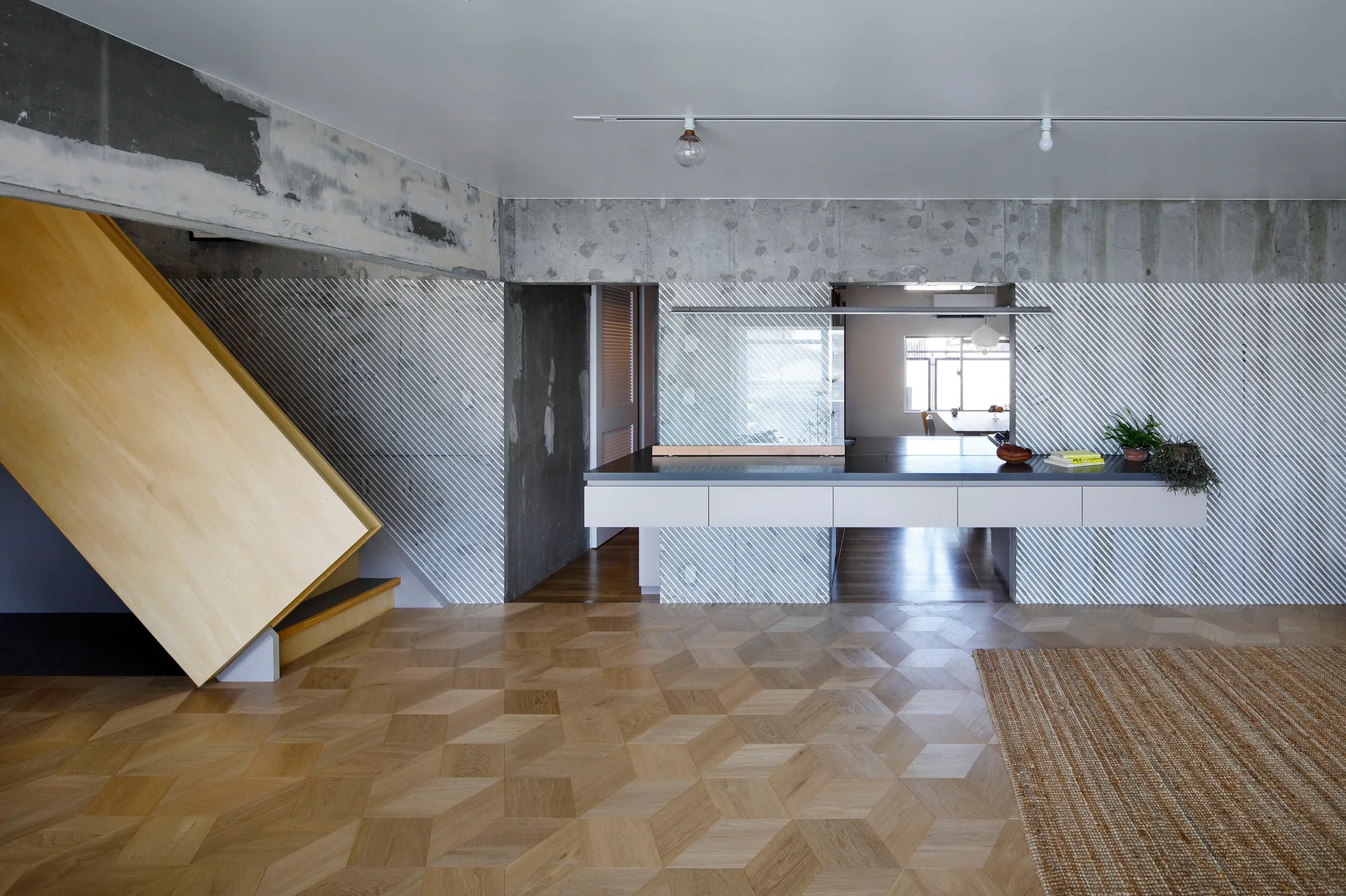 A modern kitchen space features geometric patterned wood floors, a long white floating kitchen island, diagonally striped walls, and an angled wood staircase.