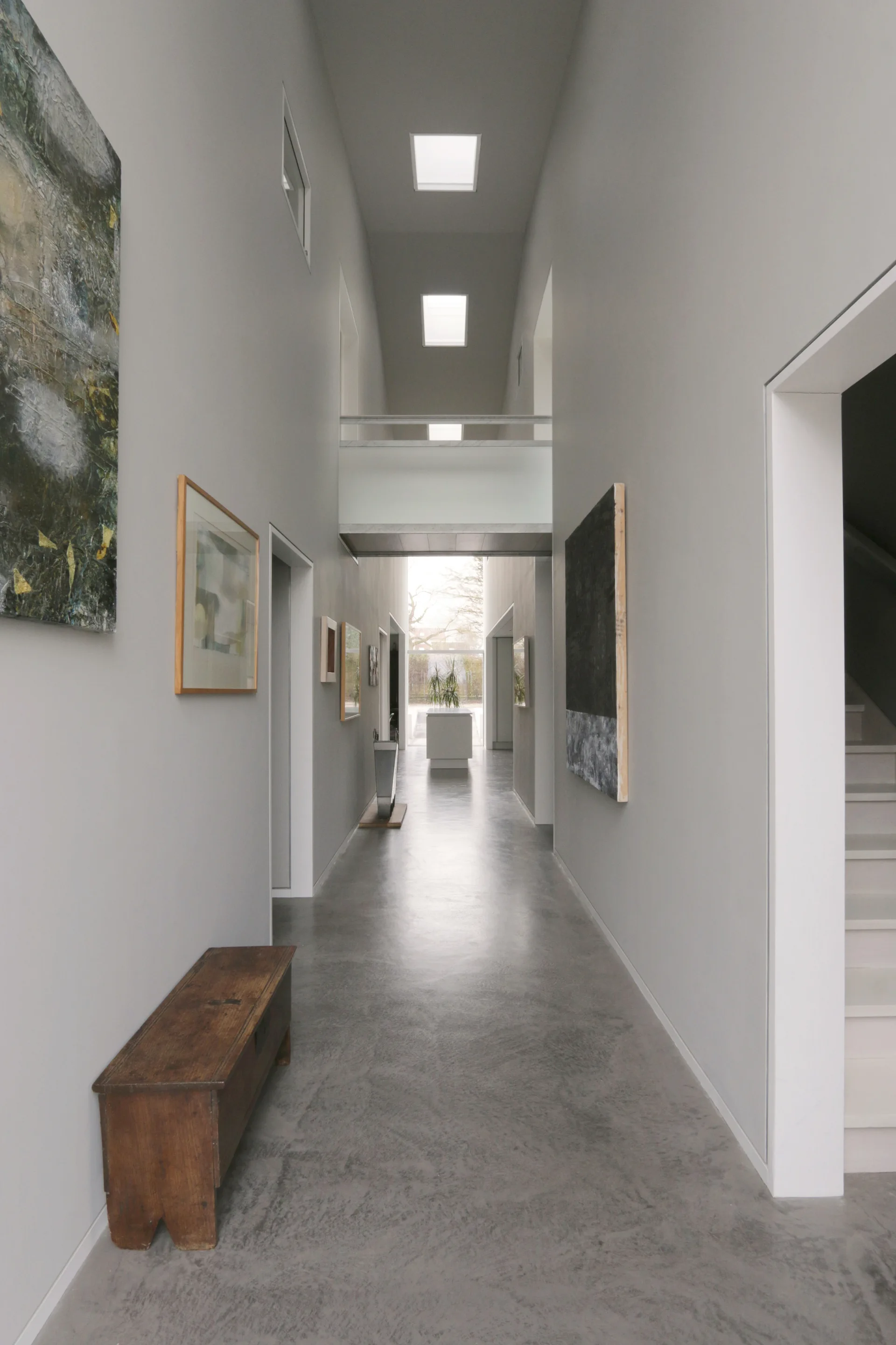A long minimalist hallway with grey walls, concrete floors, rectangular skylights, an overhead bridge, and a rustic wooden bench.