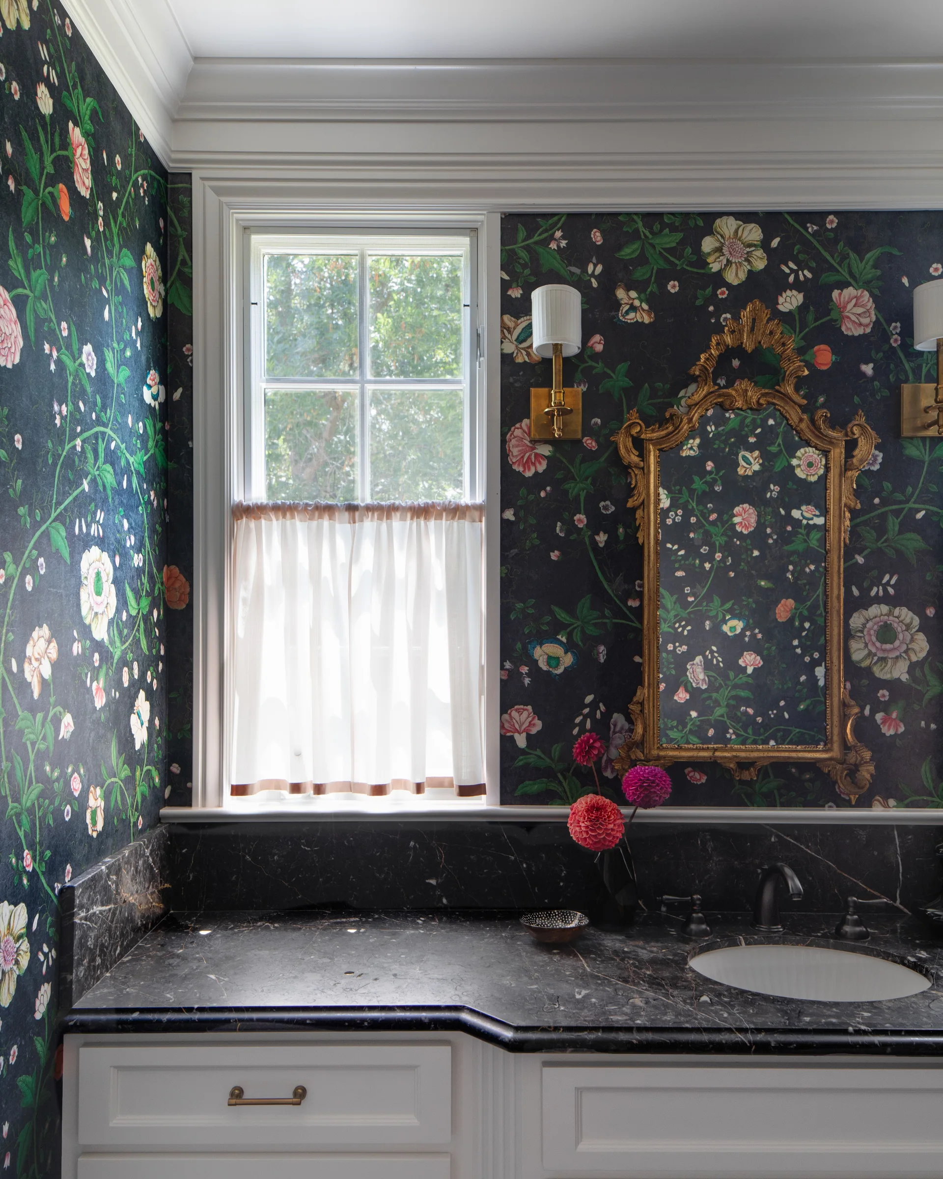 A powder room features dark floral wallpaper, an ornate gold mirror, a black marble-topped vanity, and a window with a sheer cafe curtain.