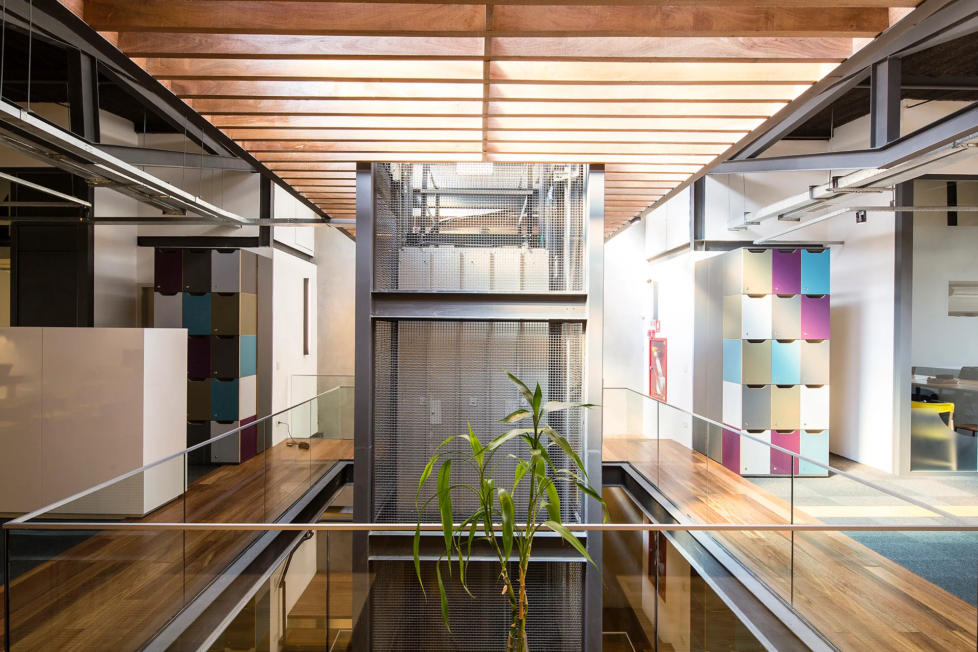 An industrial office hallway featuring a central metal mesh elevator shaft flanked by wooden walkways with glass railings under a slatted timber ceiling.