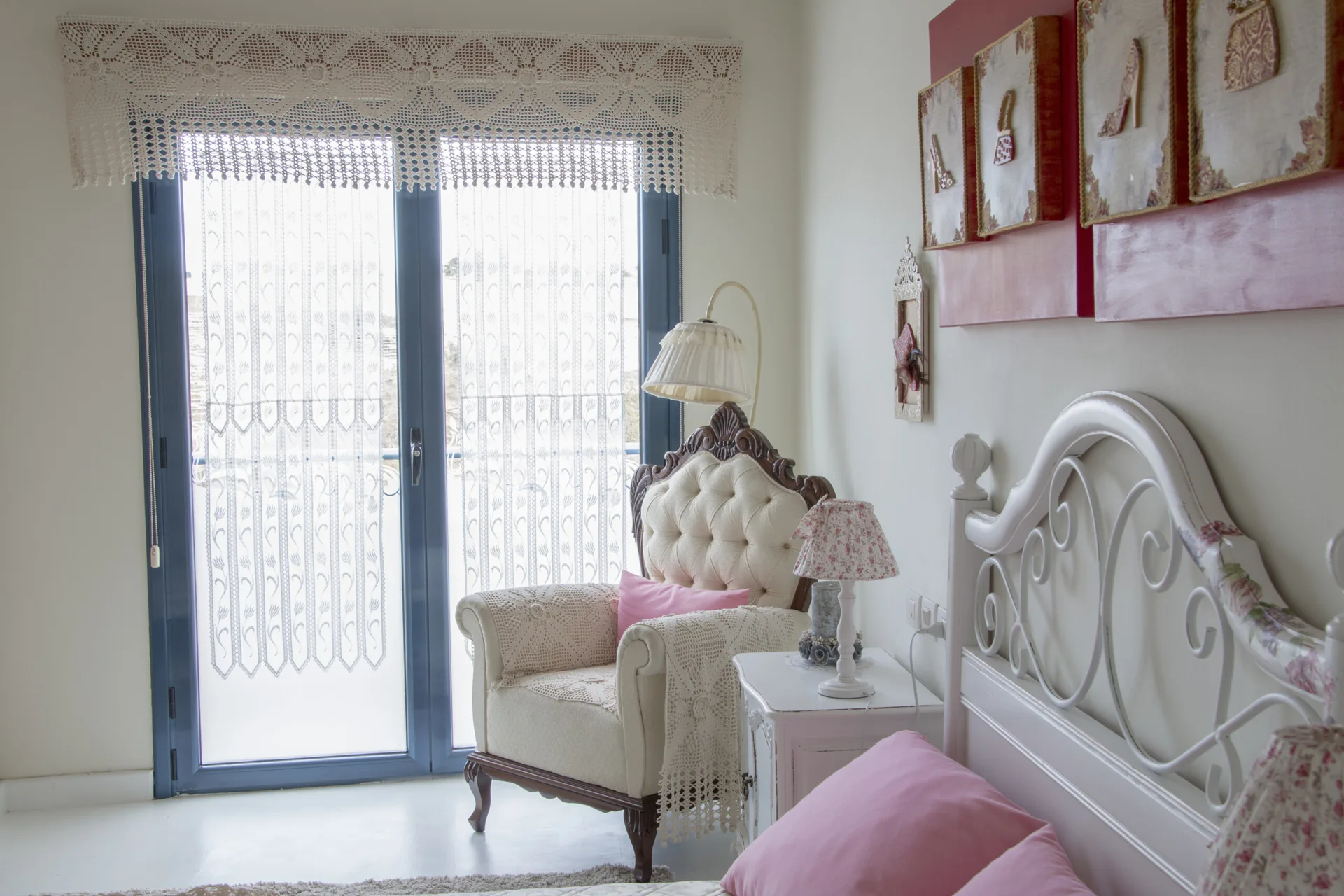 Bright bedroom corner with a white metal headboard, a tufted armchair with a crochet blanket, and glass balcony doors with lace curtains.