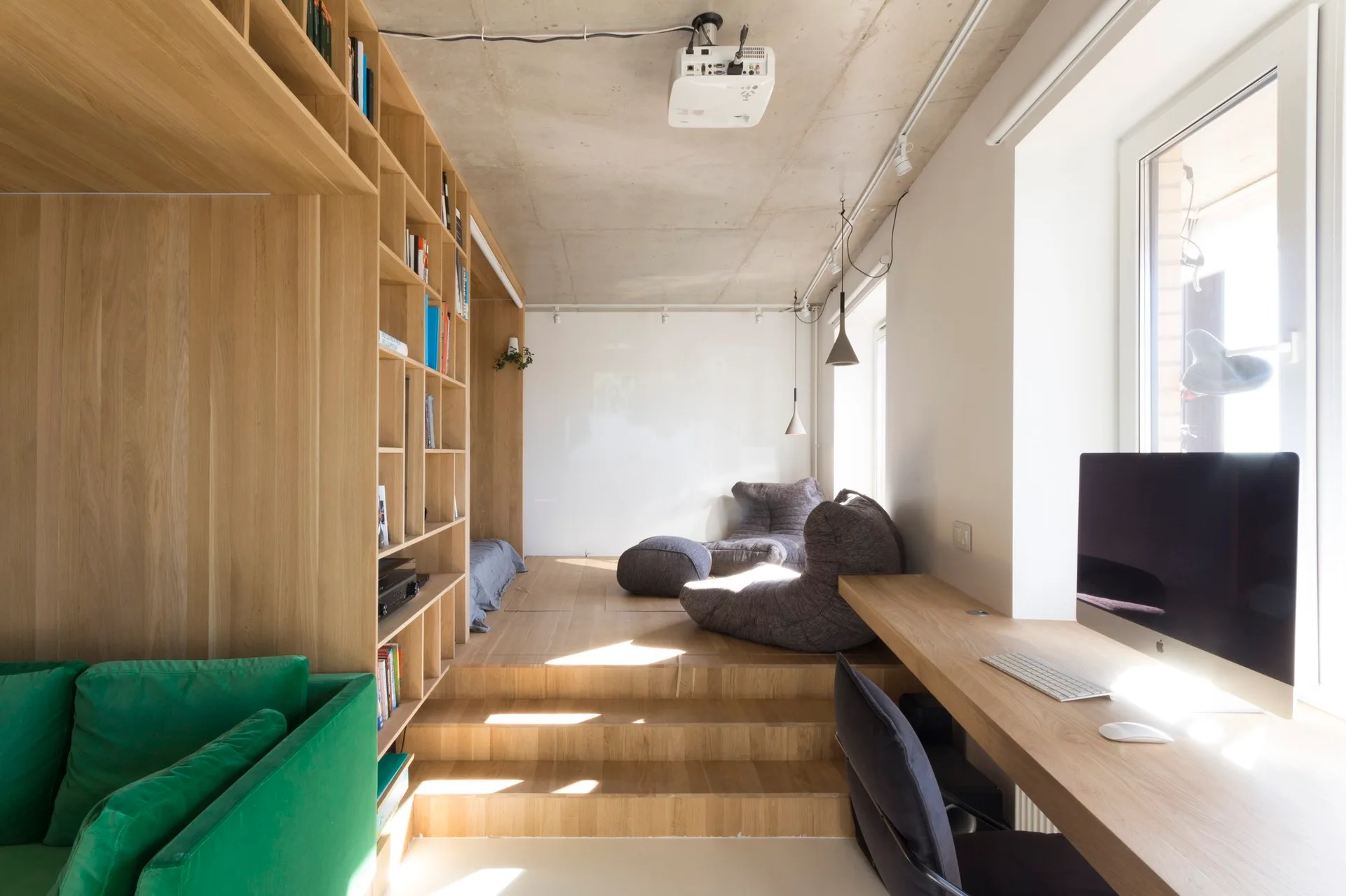 A split-level home office features a long wooden desk by the window, built-in floor-to-ceiling bookshelves, and bean bag seating on a raised platform.