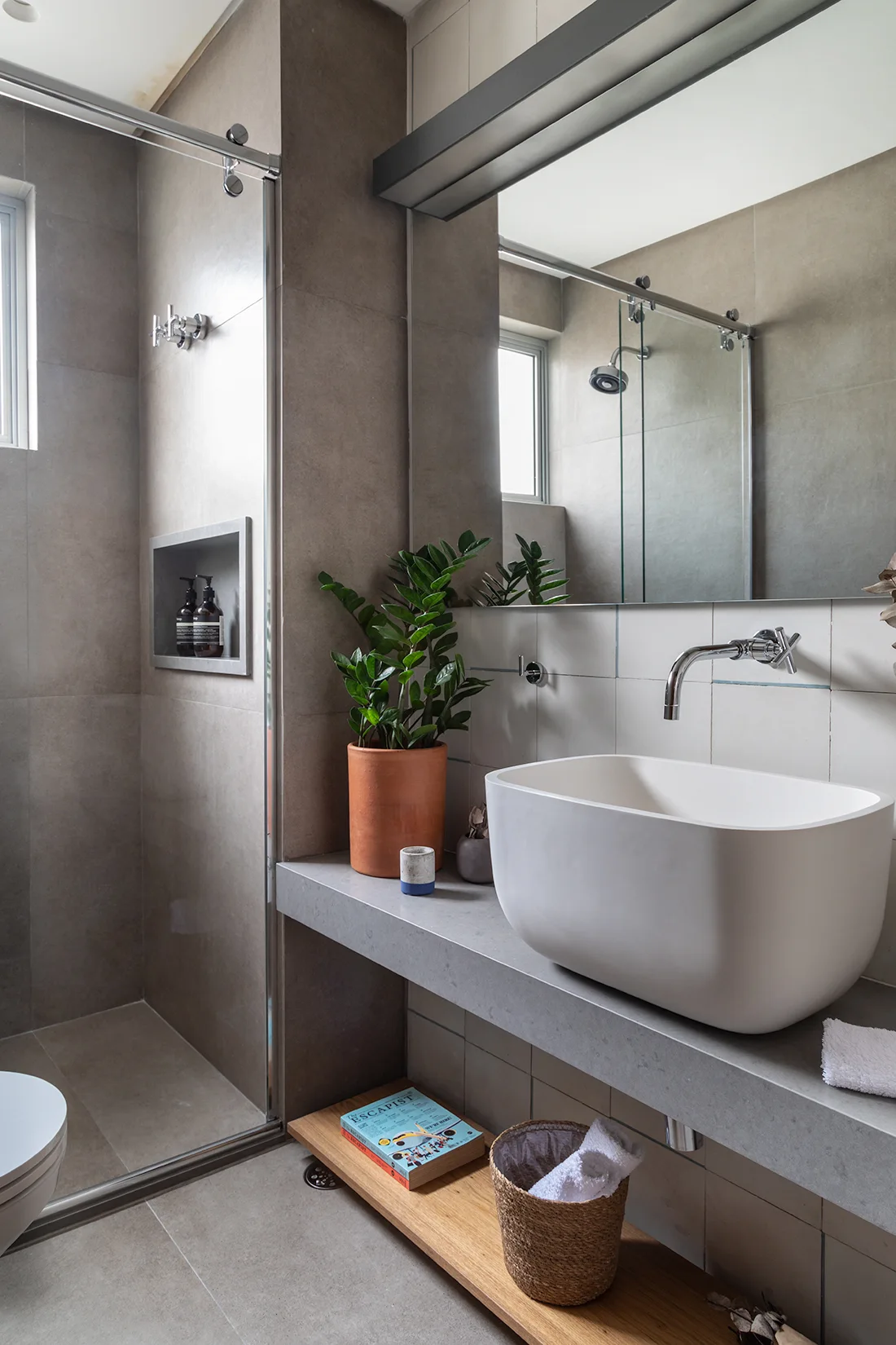 A modern bathroom features a white vessel sink on a grey stone vanity, a large mirror, and a glass-enclosed shower with grey tiled walls.