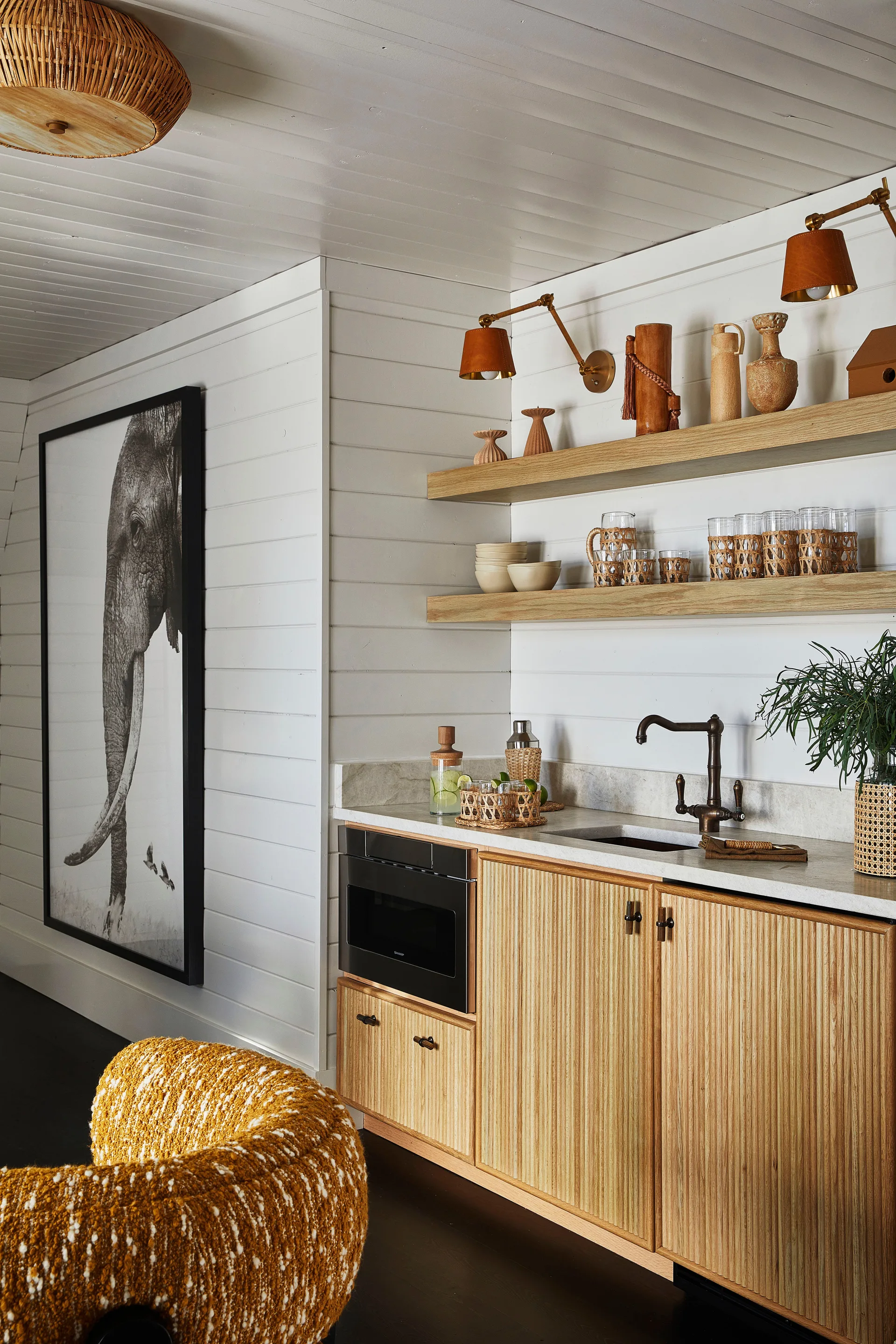 A bright kitchenette features fluted light wood cabinets, a stone countertop, white shiplap walls, and two open wooden shelves displaying decor.
