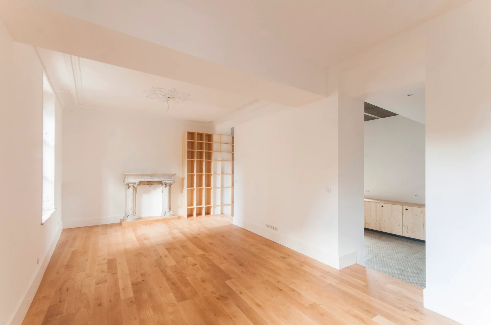 An empty white room features light wood floors, an ornate stone fireplace mantel, and a tall unfinished wooden bookshelf.
