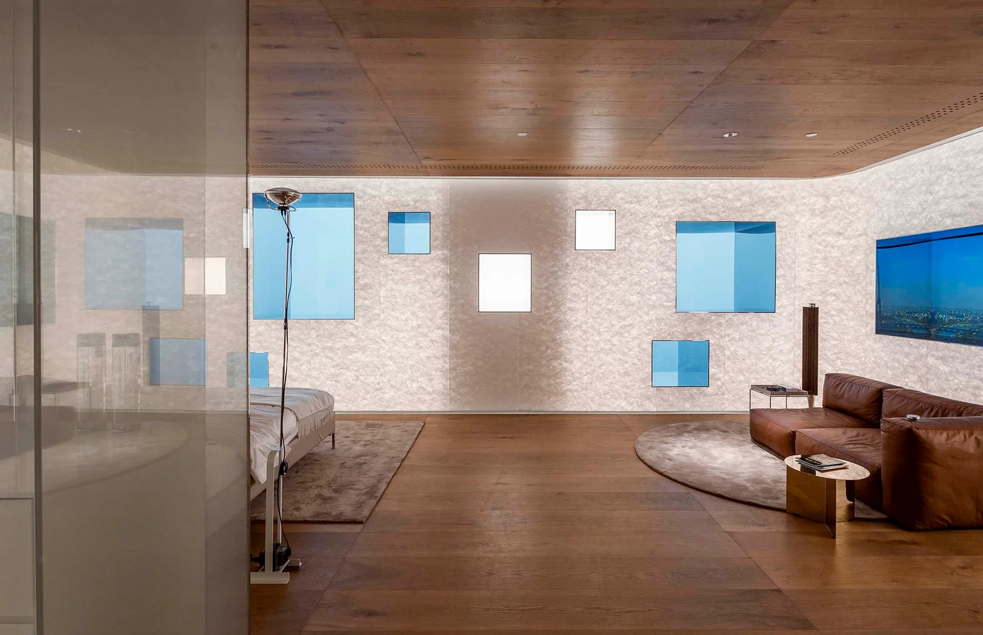 A minimalist room features a brown leather sofa, wood floors and ceiling, and a backlit textured wall inlaid with blue square windows.