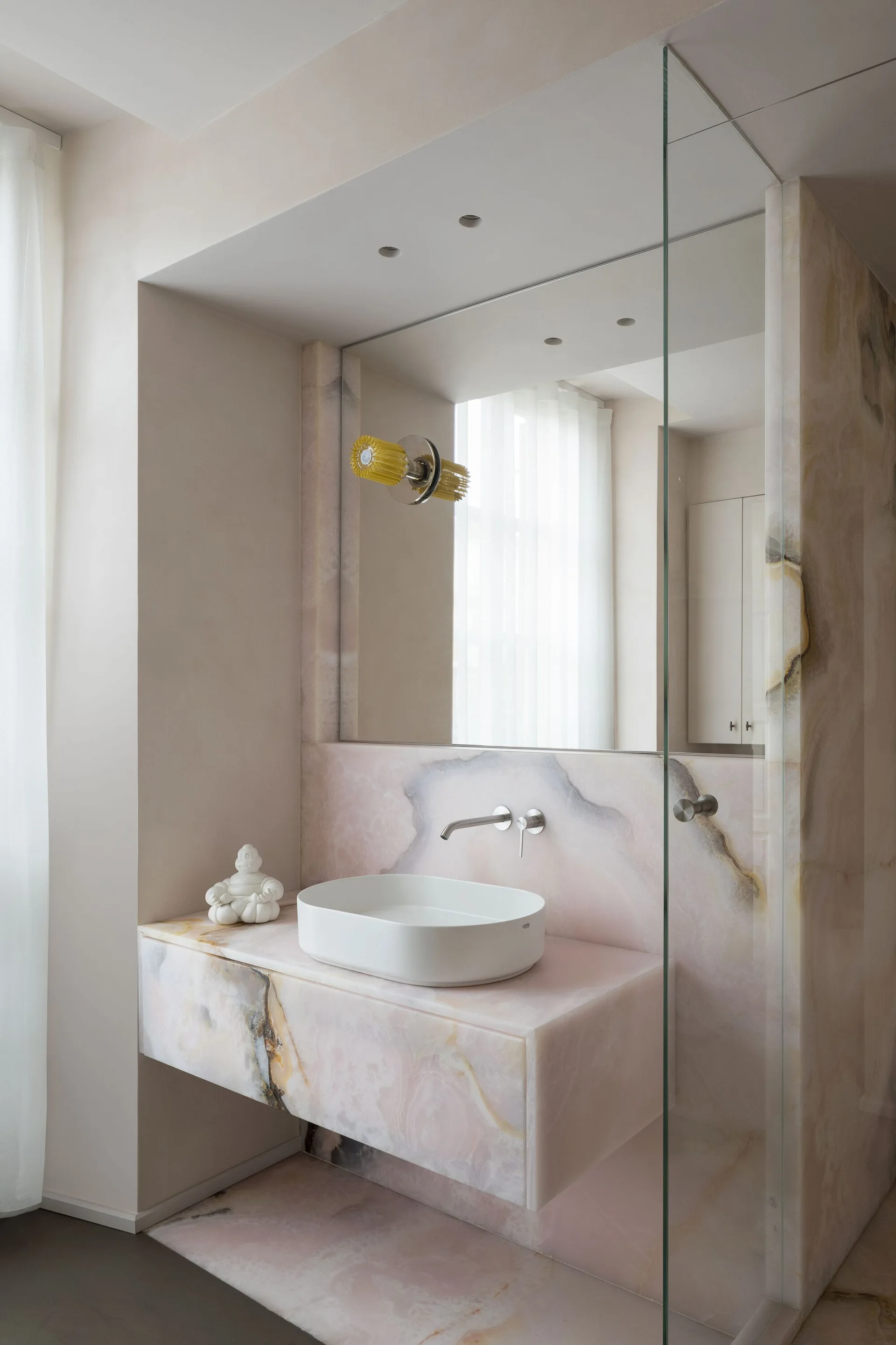 A minimalist bathroom features a floating pink stone vanity with a white vessel sink, wall-mounted faucet, and a large inset mirror.
