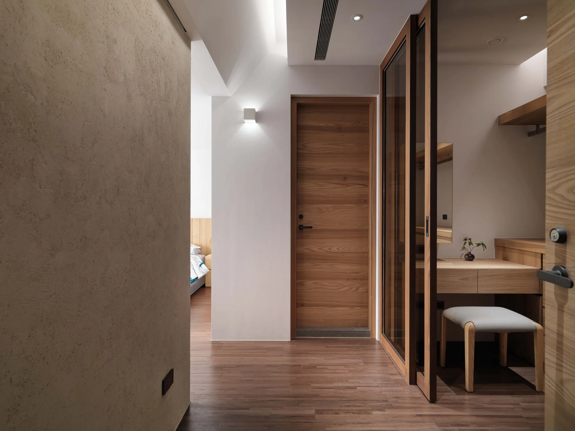 A modern hallway dressing area features a built-in wood vanity desk with a cushioned stool behind a sliding glass door partition.