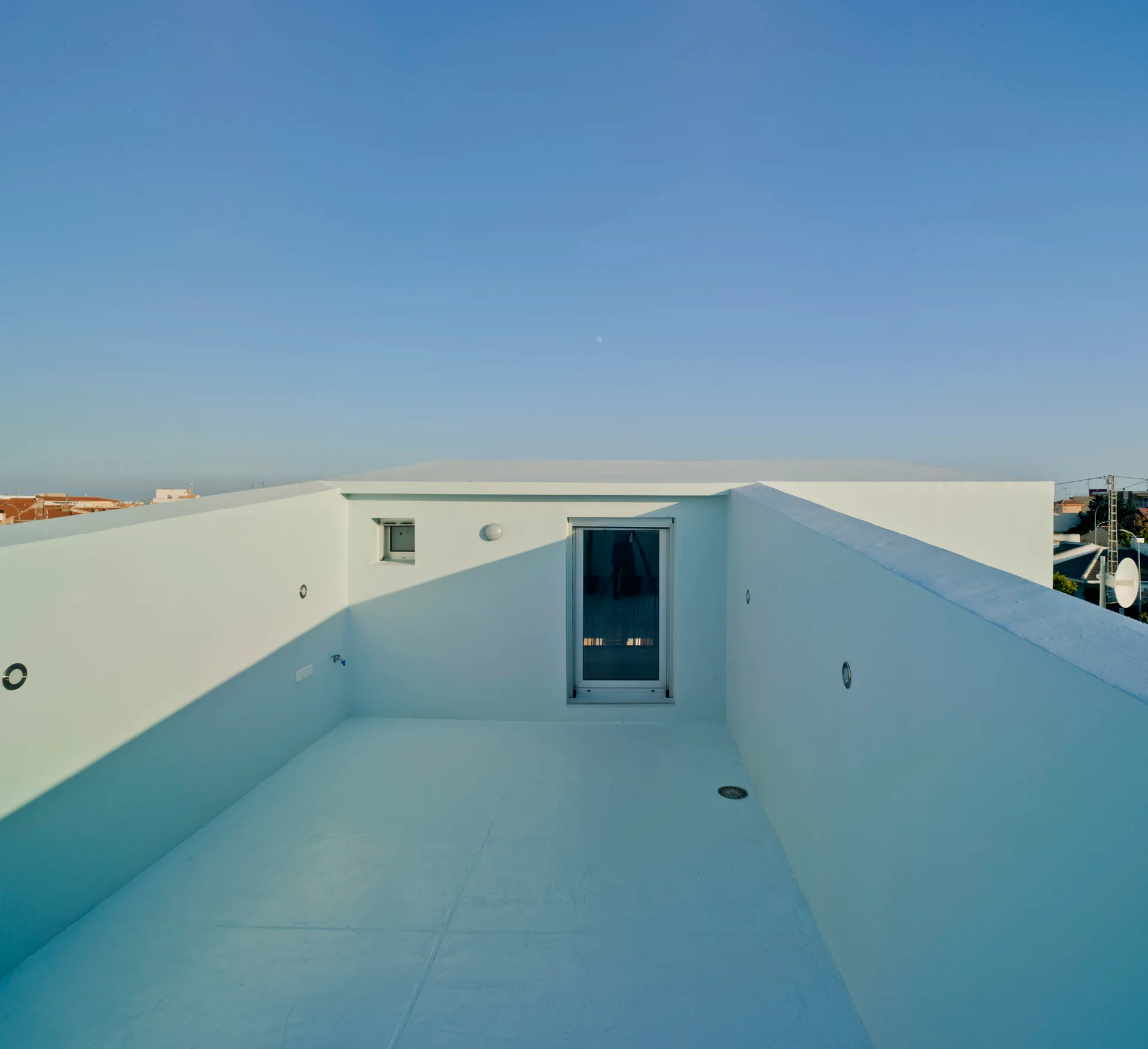 A minimalist rooftop terrace with pale blue walls and flooring features a central glass door and a small square window under a clear sky.