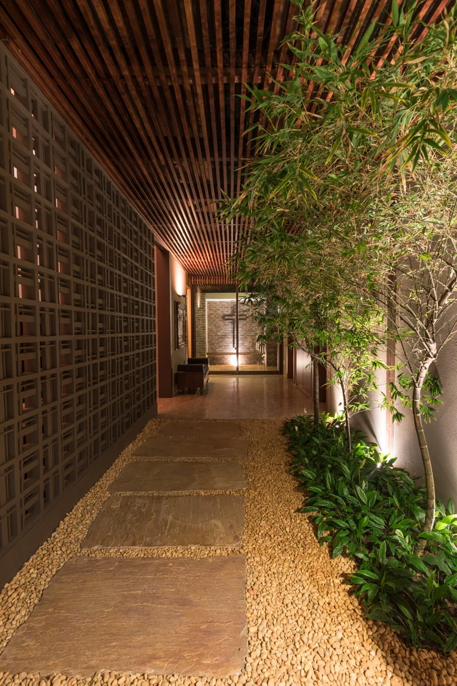 An indoor corridor featuring a slatted wood ceiling, a patterned breeze block wall, and a stepping stone path set in pebbles next to bamboo plants.