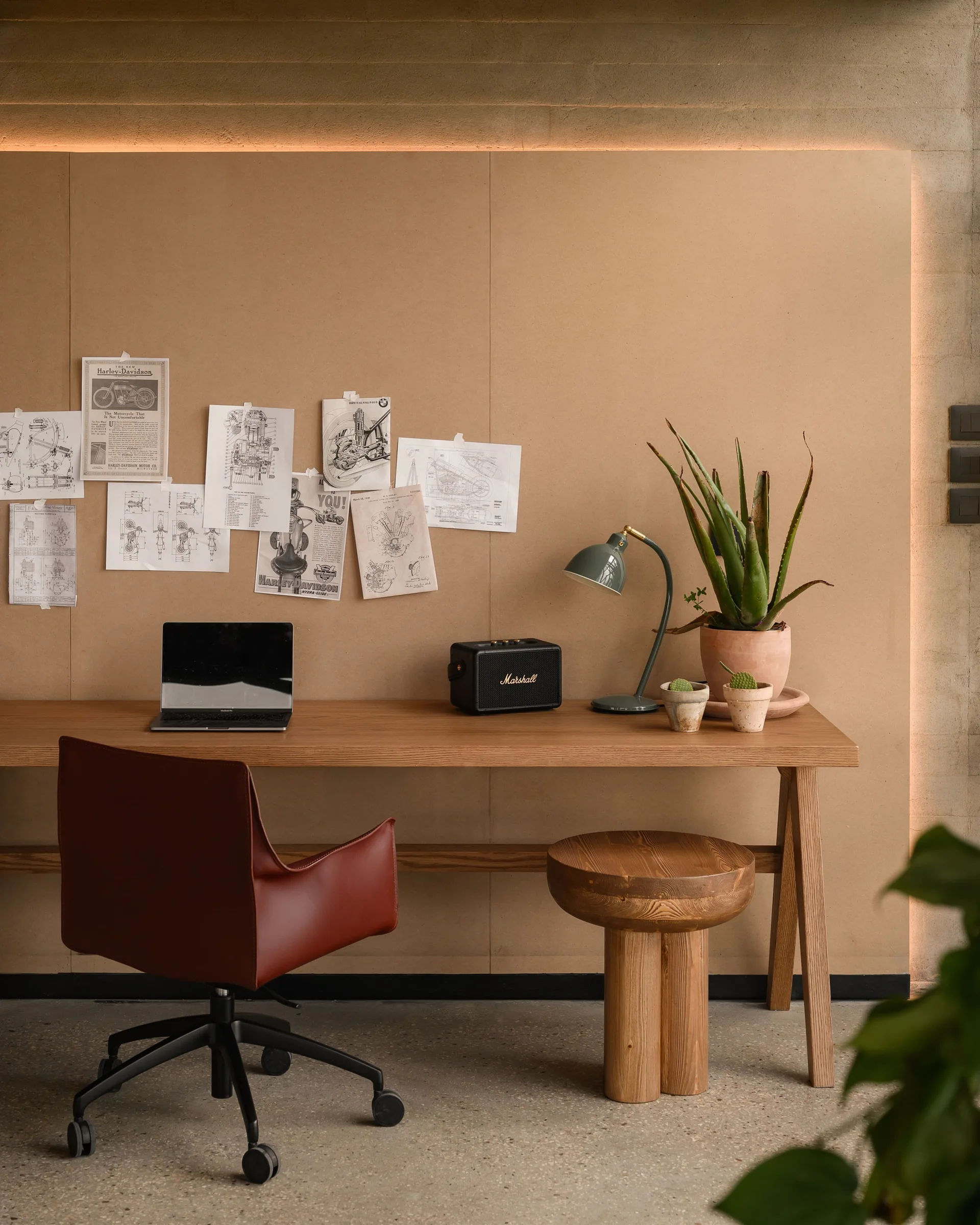 A minimalist home office workspace features a long wooden desk, a maroon desk chair, and technical drawings taped to a backlit wall panel.