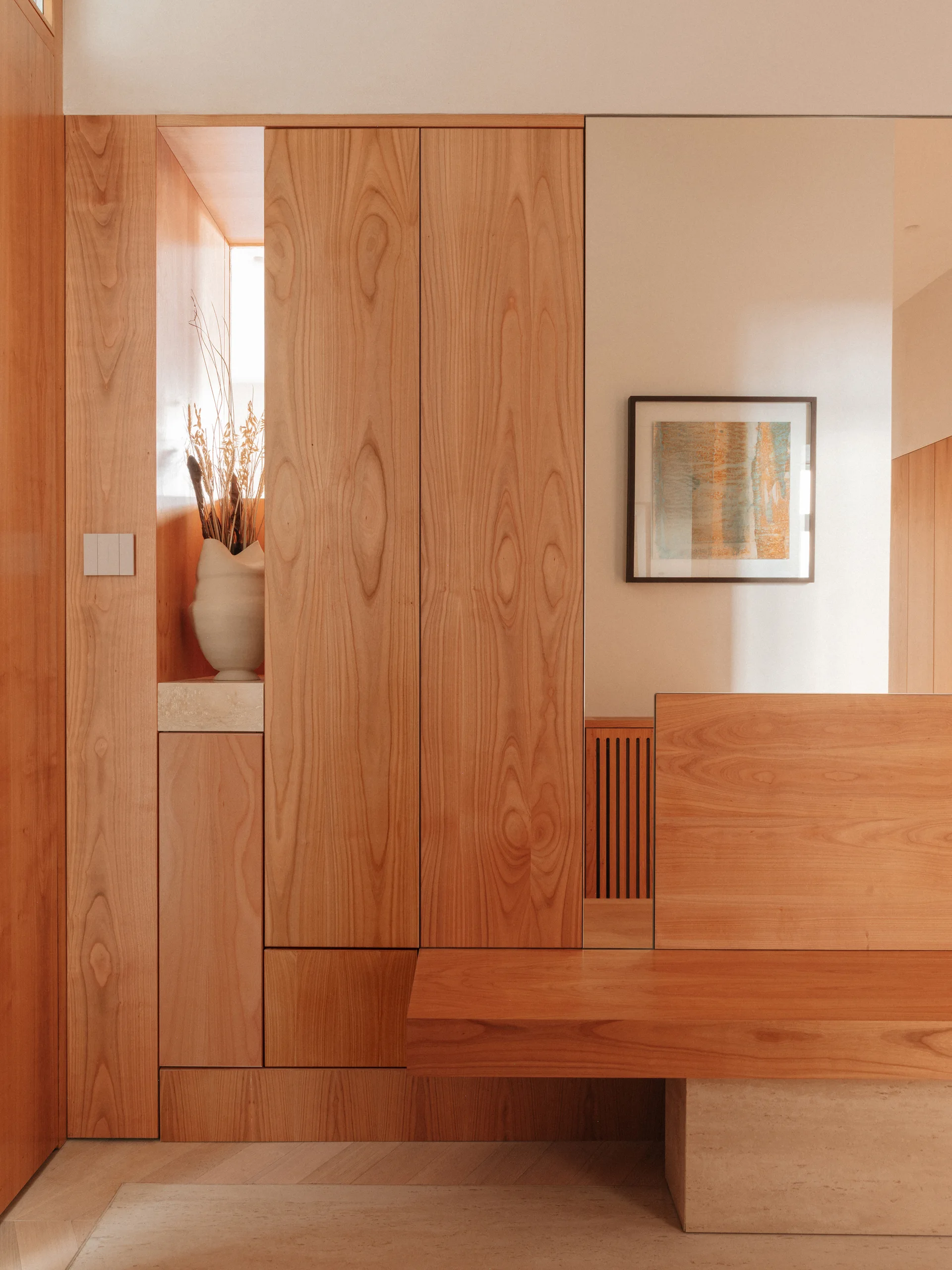 Built-in wood cabinetry lines an entryway featuring a recessed display niche with a white vase and a low wooden bench resting on a stone base.