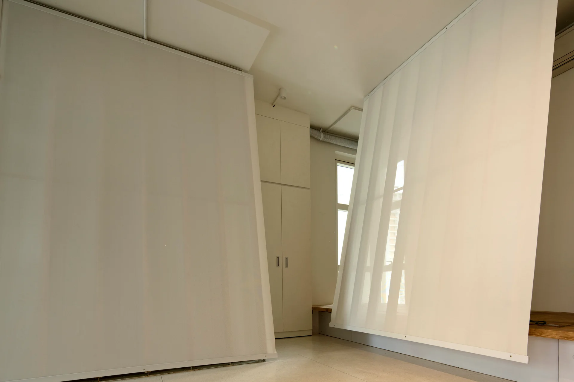 Translucent white fabric panels suspended from ceiling tracks act as room dividers near built-in cabinetry in a minimalist room.