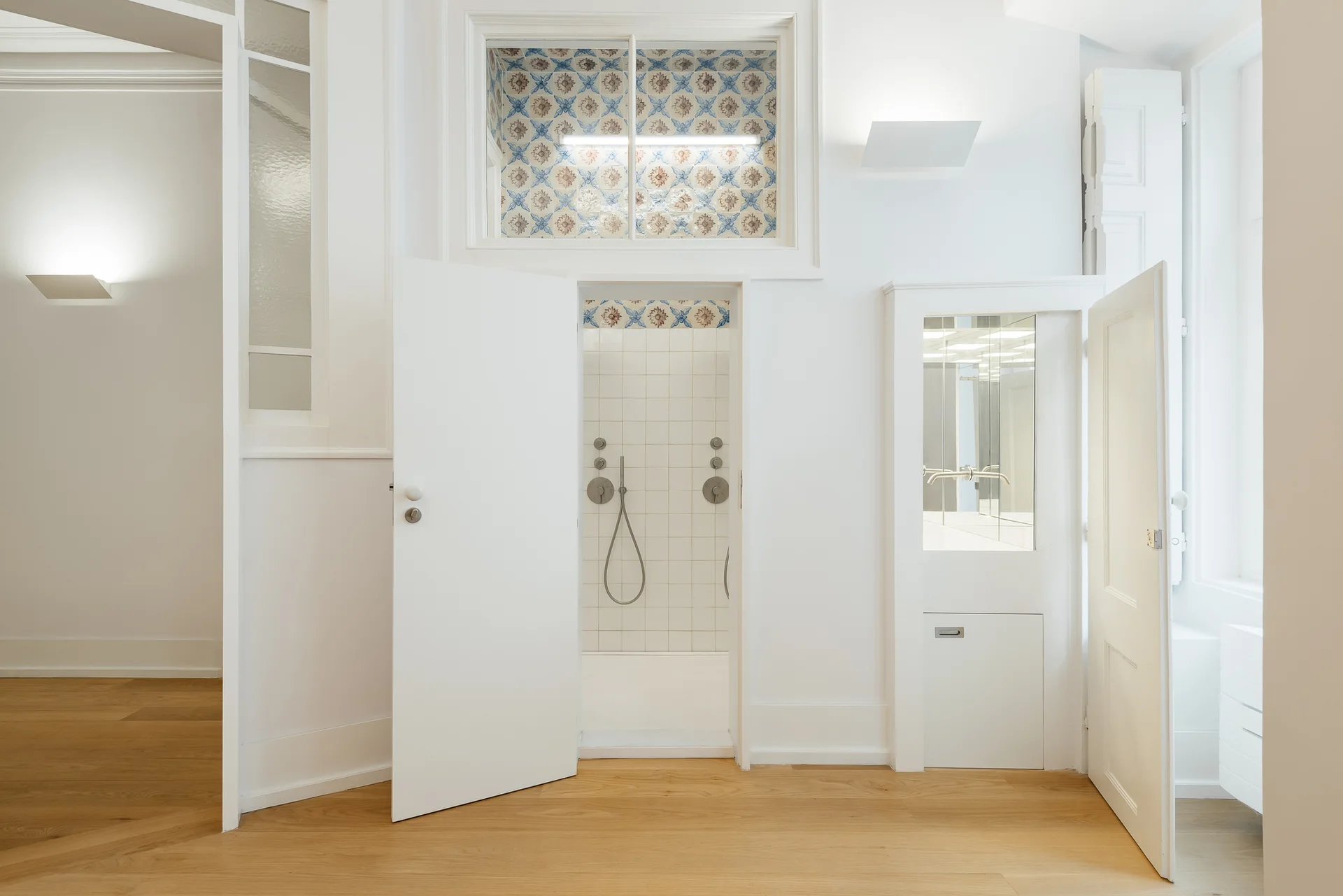 A bright white bathroom features a tile-lined shower, custom built-in vanity with a mirror, natural wood floors, and a patterned transom window.