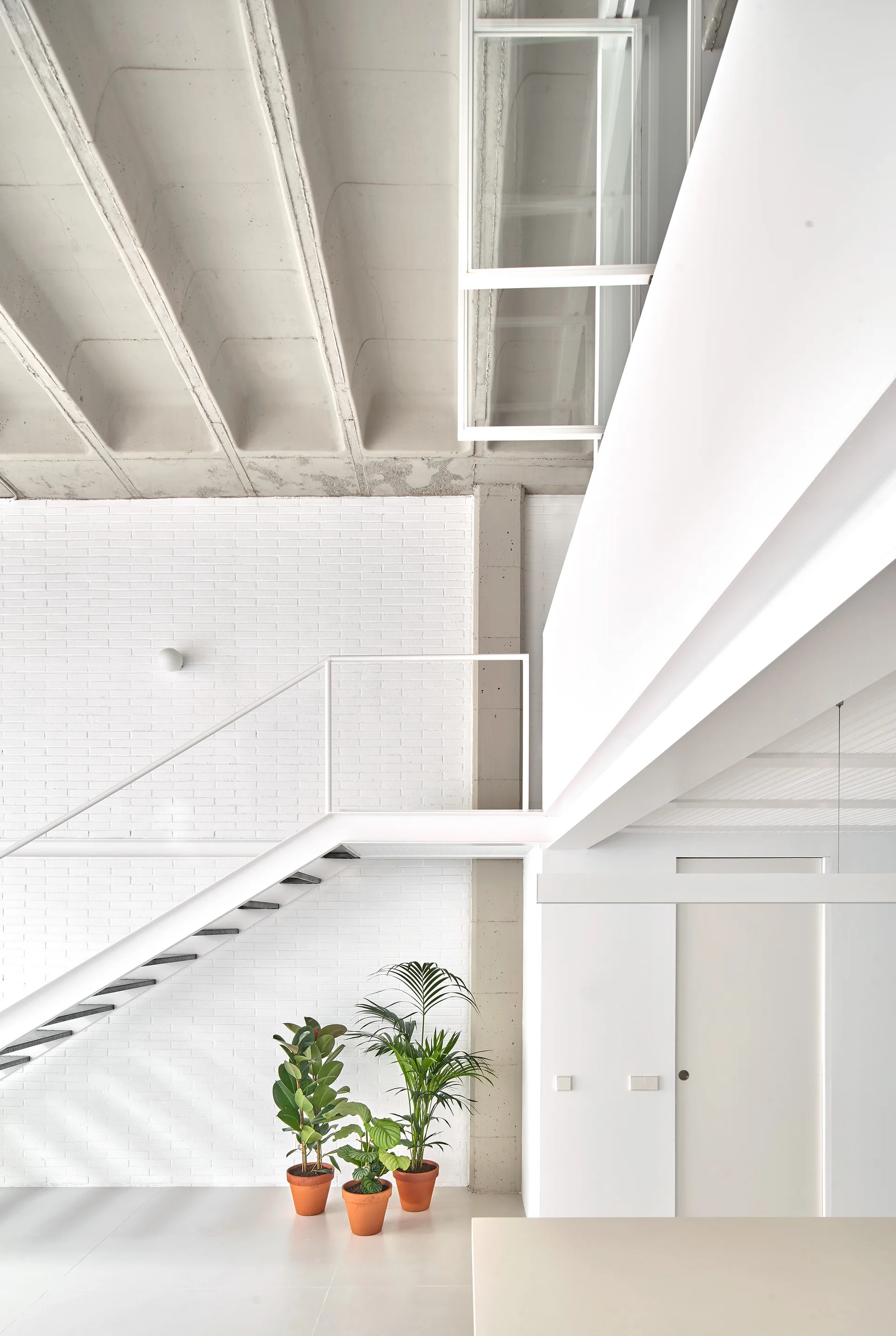 A white metal staircase rises against a painted brick wall under an exposed concrete ceiling with three terracotta potted plants at the base.