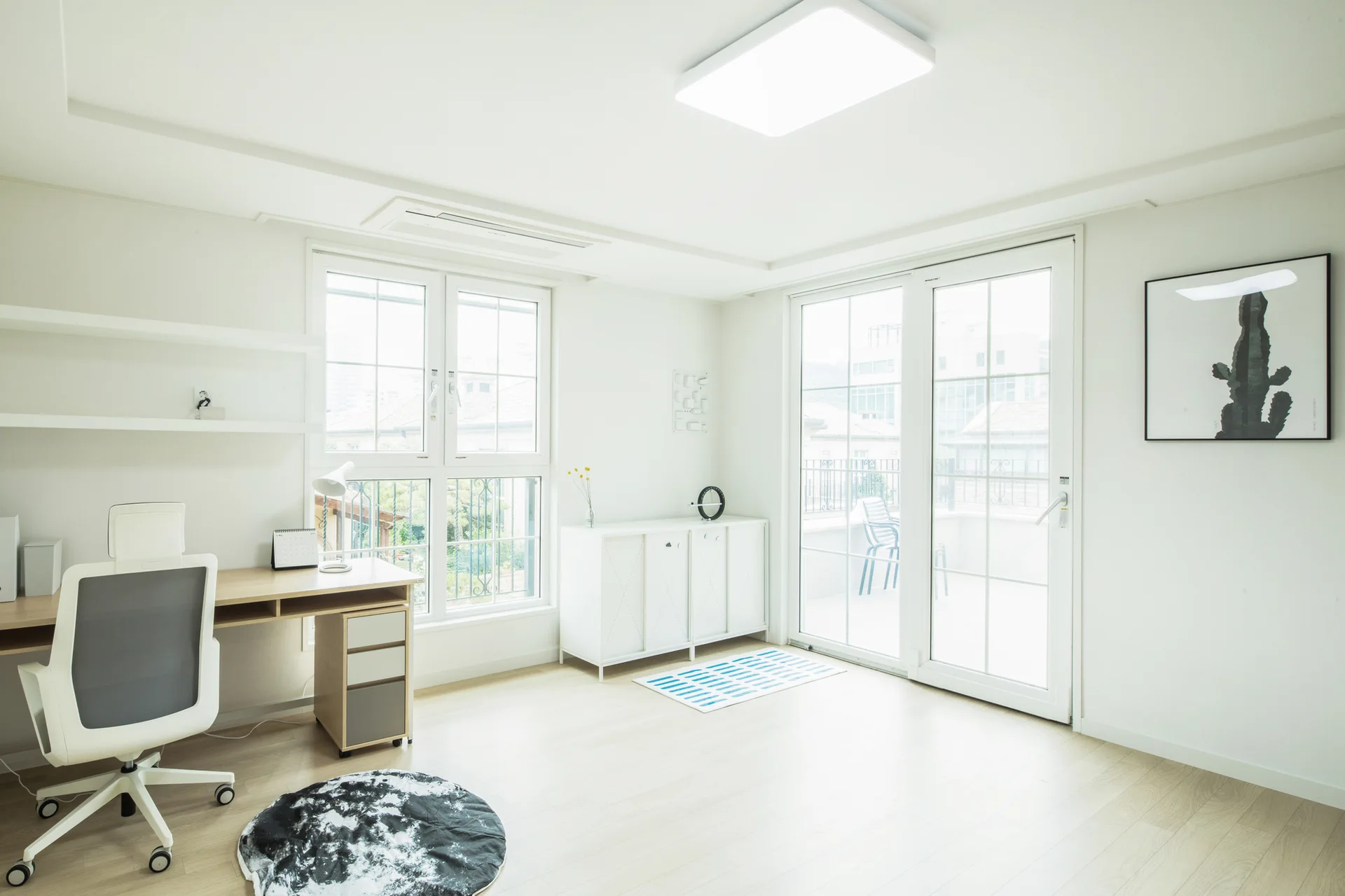A bright home office features a light wood desk, a white mesh office chair, a white sideboard, and large glass windows leading to a balcony.
