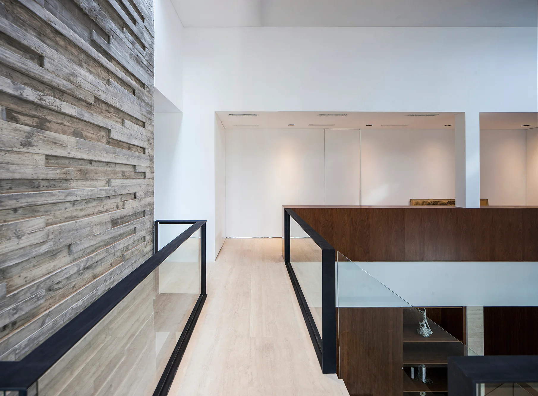 A modern hallway landing features a textured wood plank feature wall, glass railings with black frames, and a light stone floor walkway.