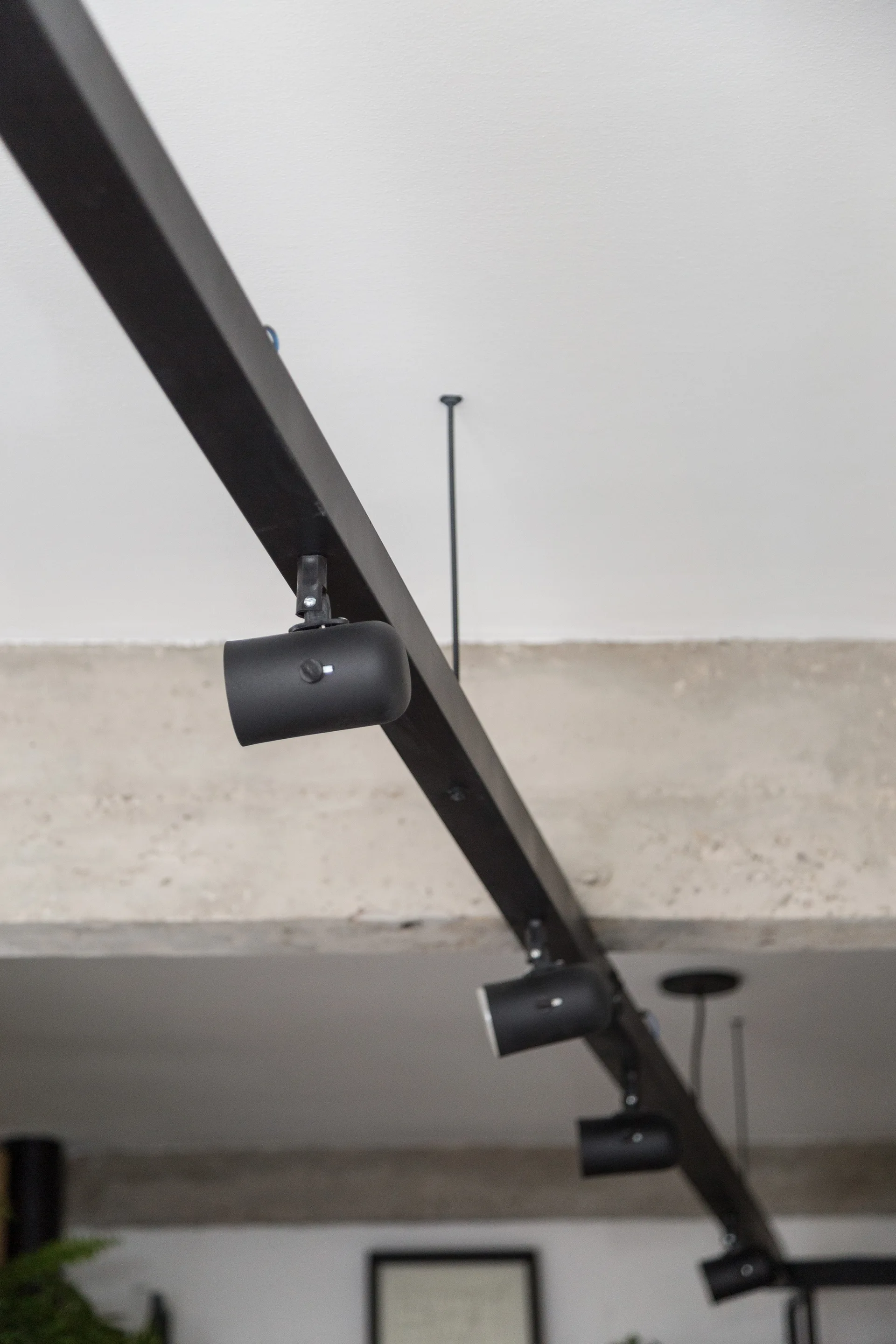A close-up of a matte black track lighting fixture with cylindrical spotlight heads mounted on a white ceiling near a textured concrete beam.