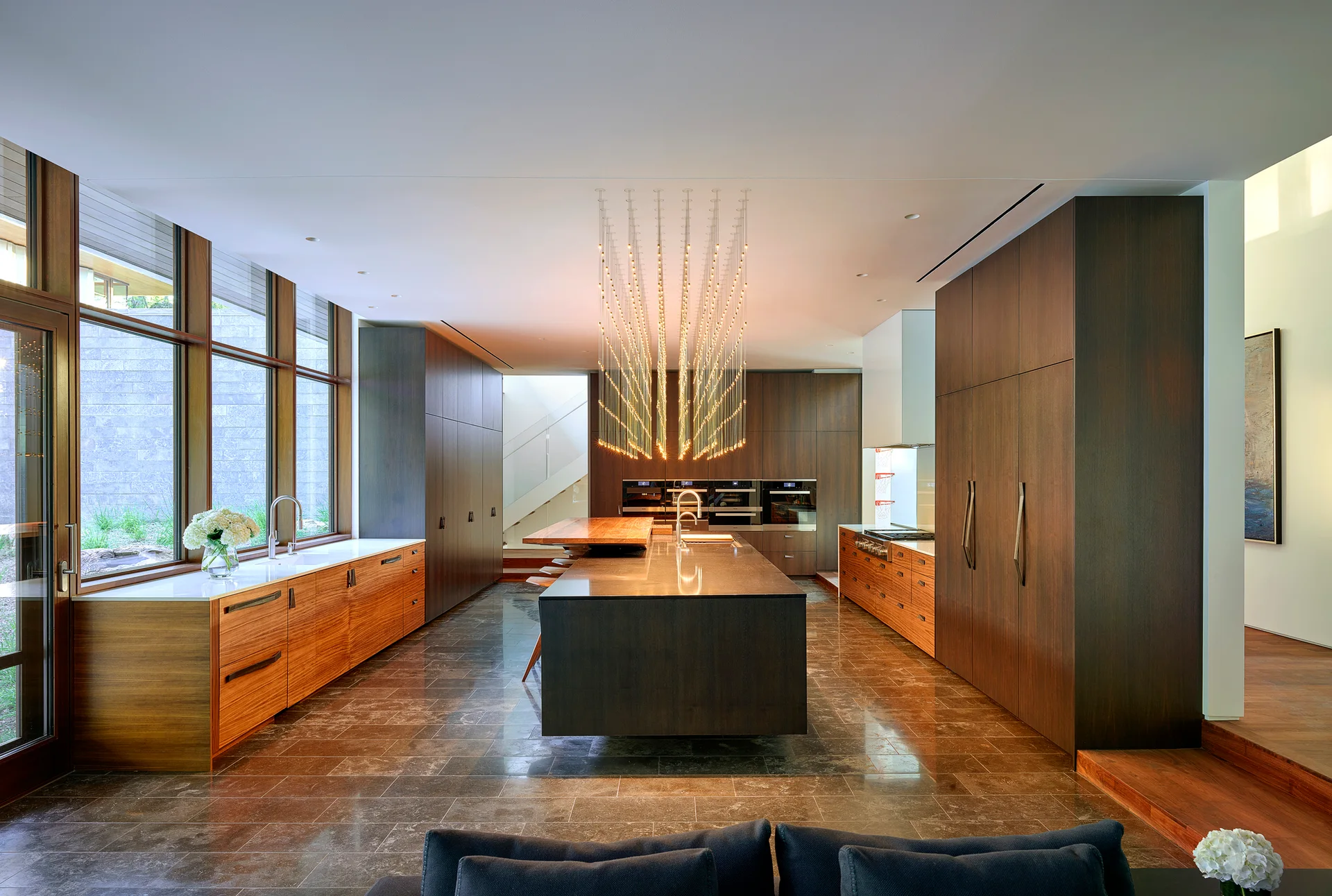 A large modern kitchen features dark wood floor-to-ceiling cabinets, a central island, stone tile flooring, and a cascading multi-strand light fixture.