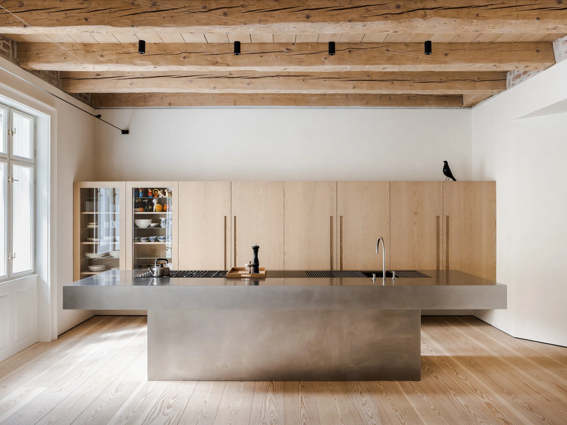 A minimalist kitchen features a large stainless steel island, light wood cabinetry, and exposed wooden ceiling beams above light wood floors.