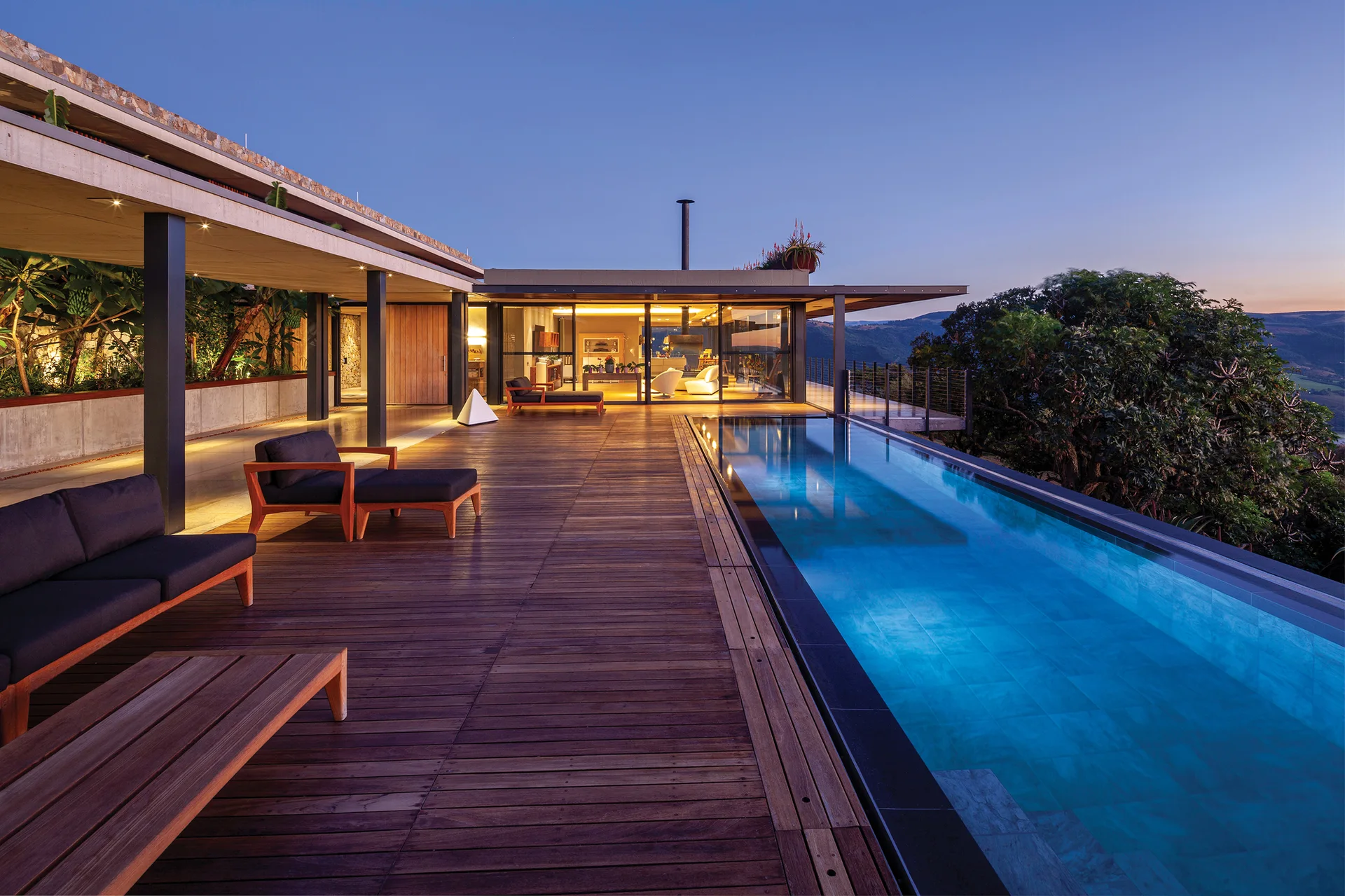 A long lap pool illuminated with blue light sits on a wide wooden deck next to a modern home with sliding glass doors at dusk.