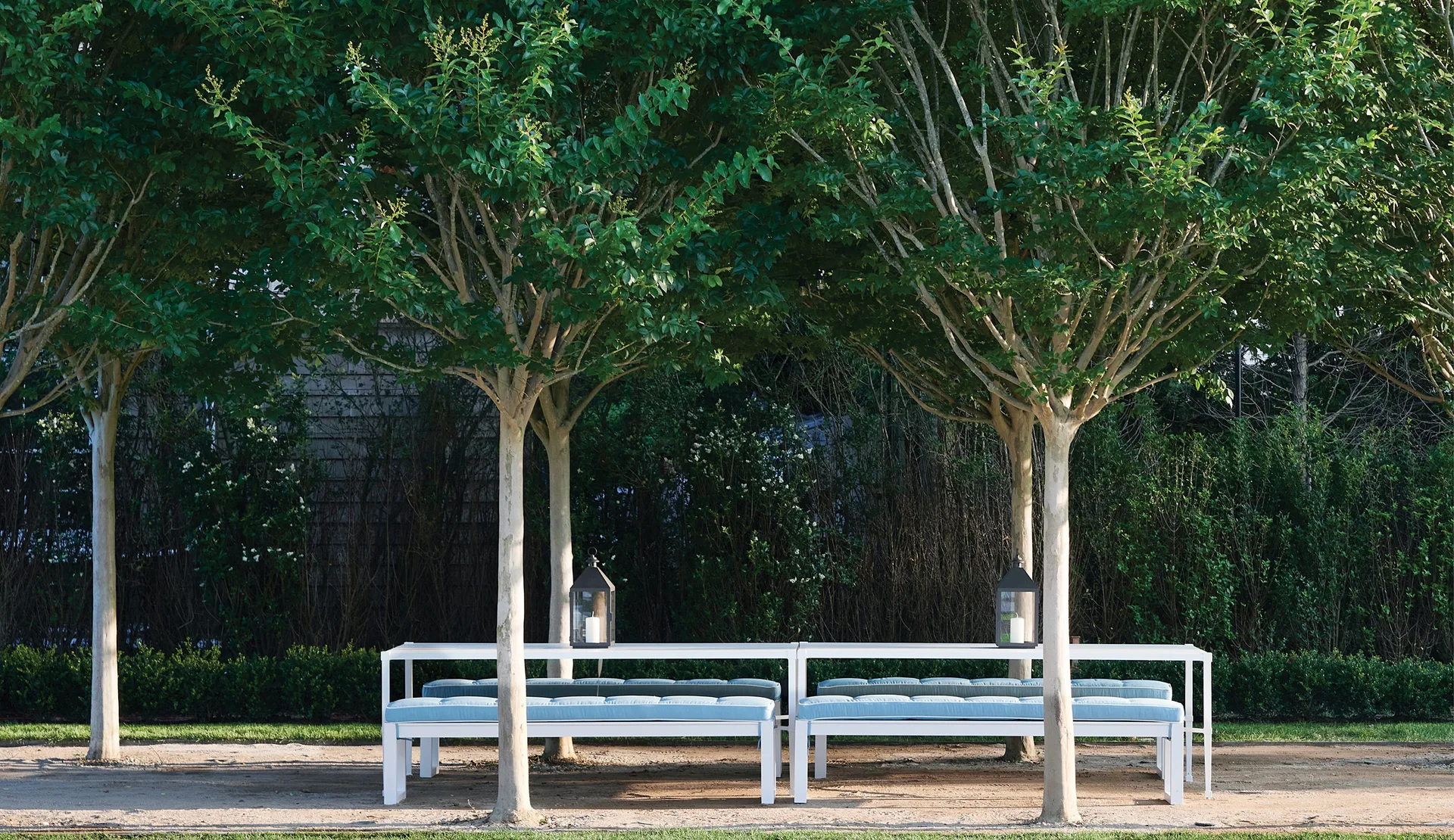 A long white metal dining table and benches with light blue cushions are arranged on a gravel path under a row of manicured trees.