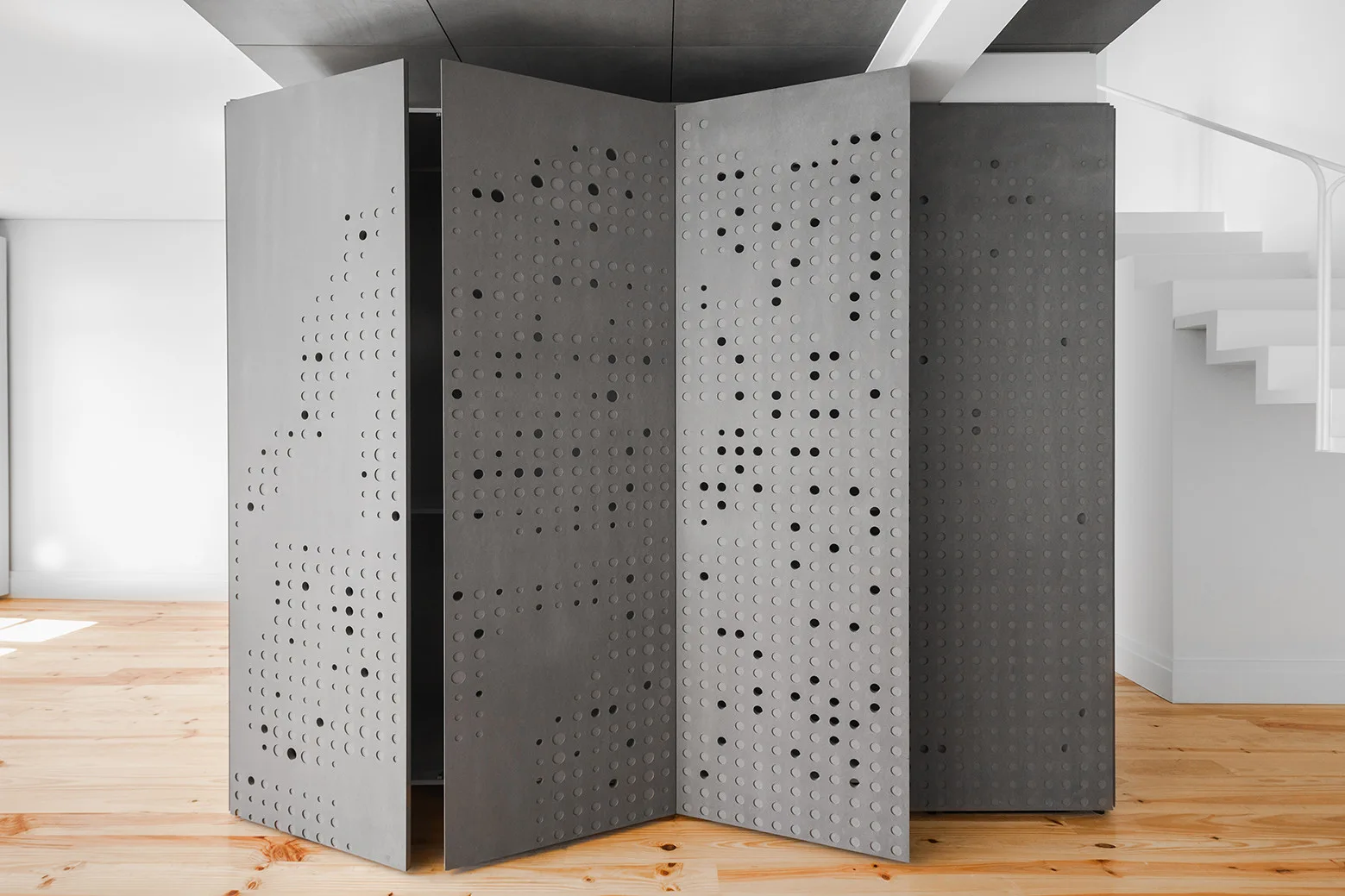 A tall grey folding screen with circular perforations stands on a light wood floor in a minimalist room next to a white staircase.