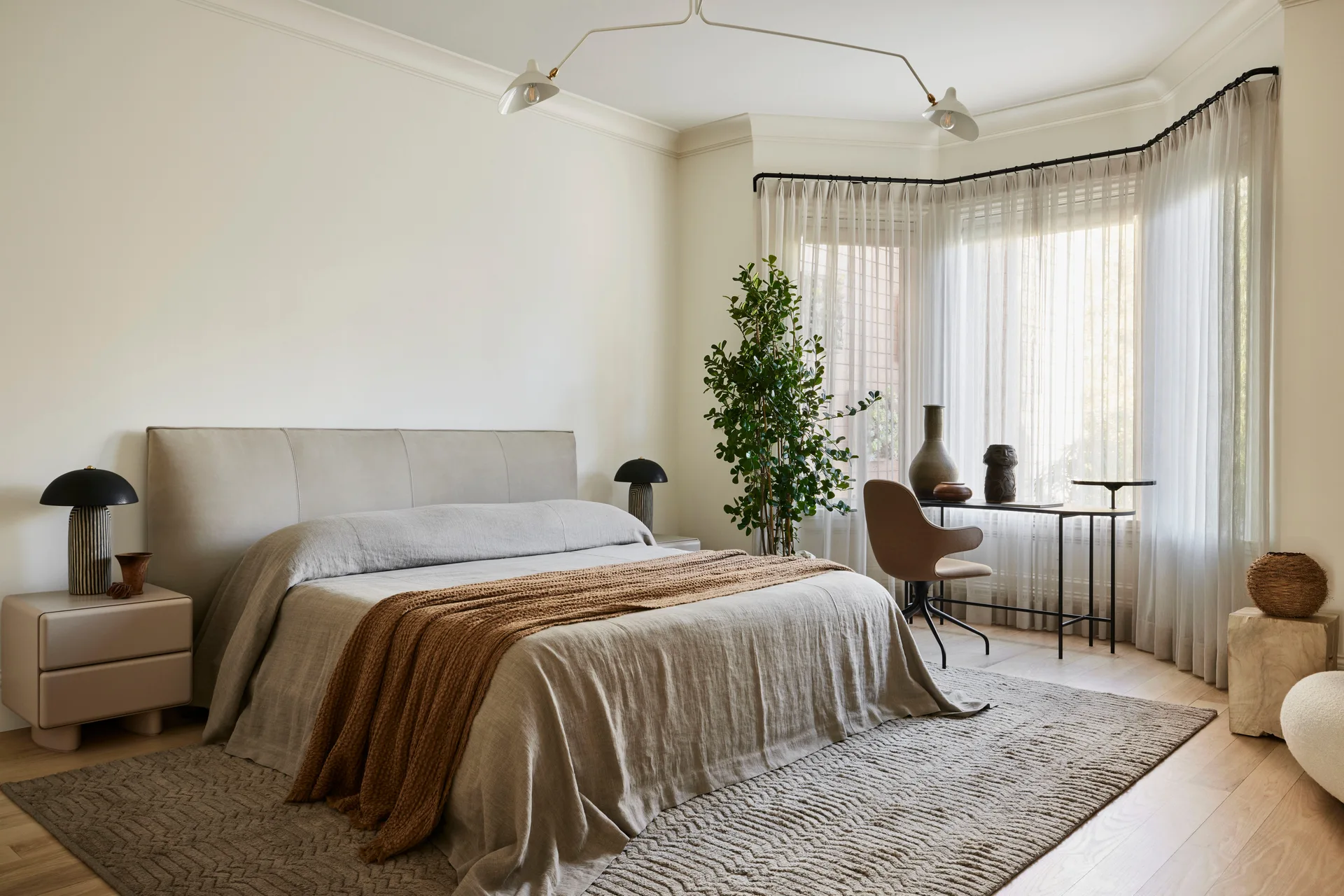A neutral-toned bedroom features a bed with beige linen bedding, a textured rug, two mushroom lamps, and a slim desk set in a curved bay window.