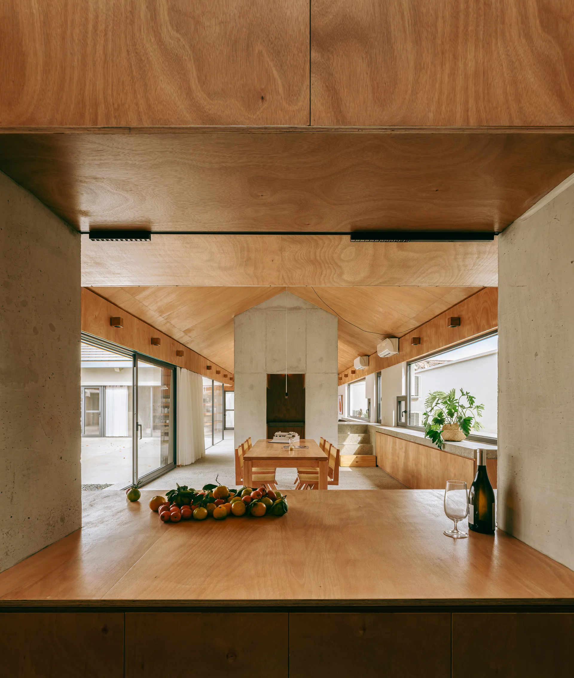 Viewed through a concrete frame, a minimalist dining room features a vaulted plywood ceiling, a wooden dining set, and large glass doors.