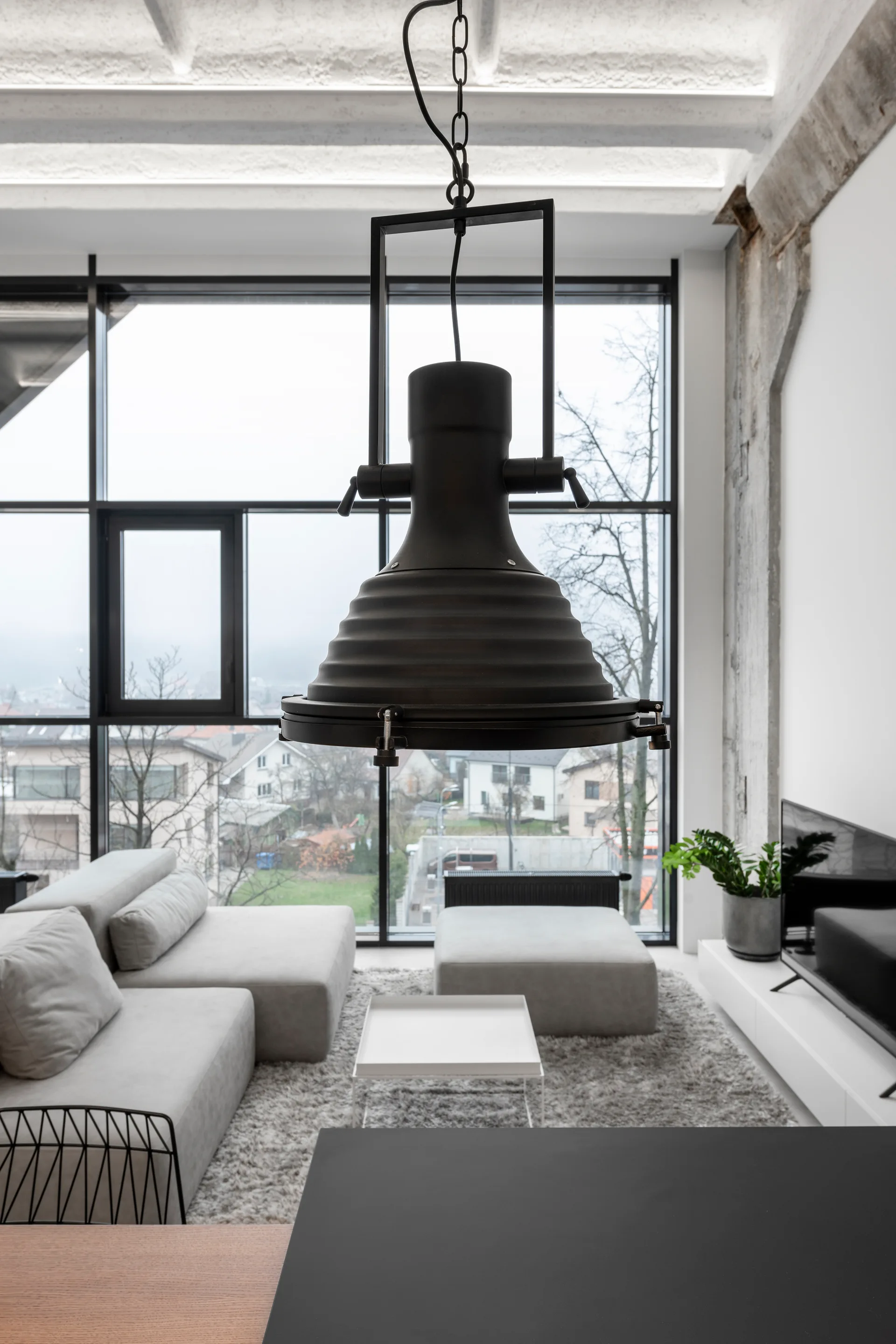 A minimalist loft living room features a grey sectional sofa, a large black industrial pendant light, and a massive floor-to-ceiling grid window.