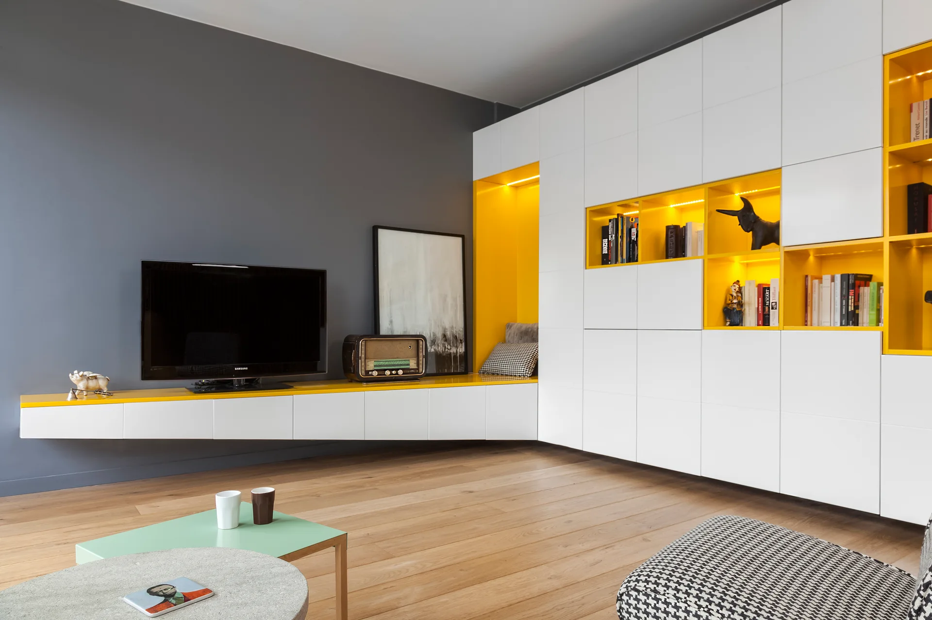 A modern living room features a dark gray wall with a floating console below a TV, next to white built-in cabinets with illuminated yellow shelves.