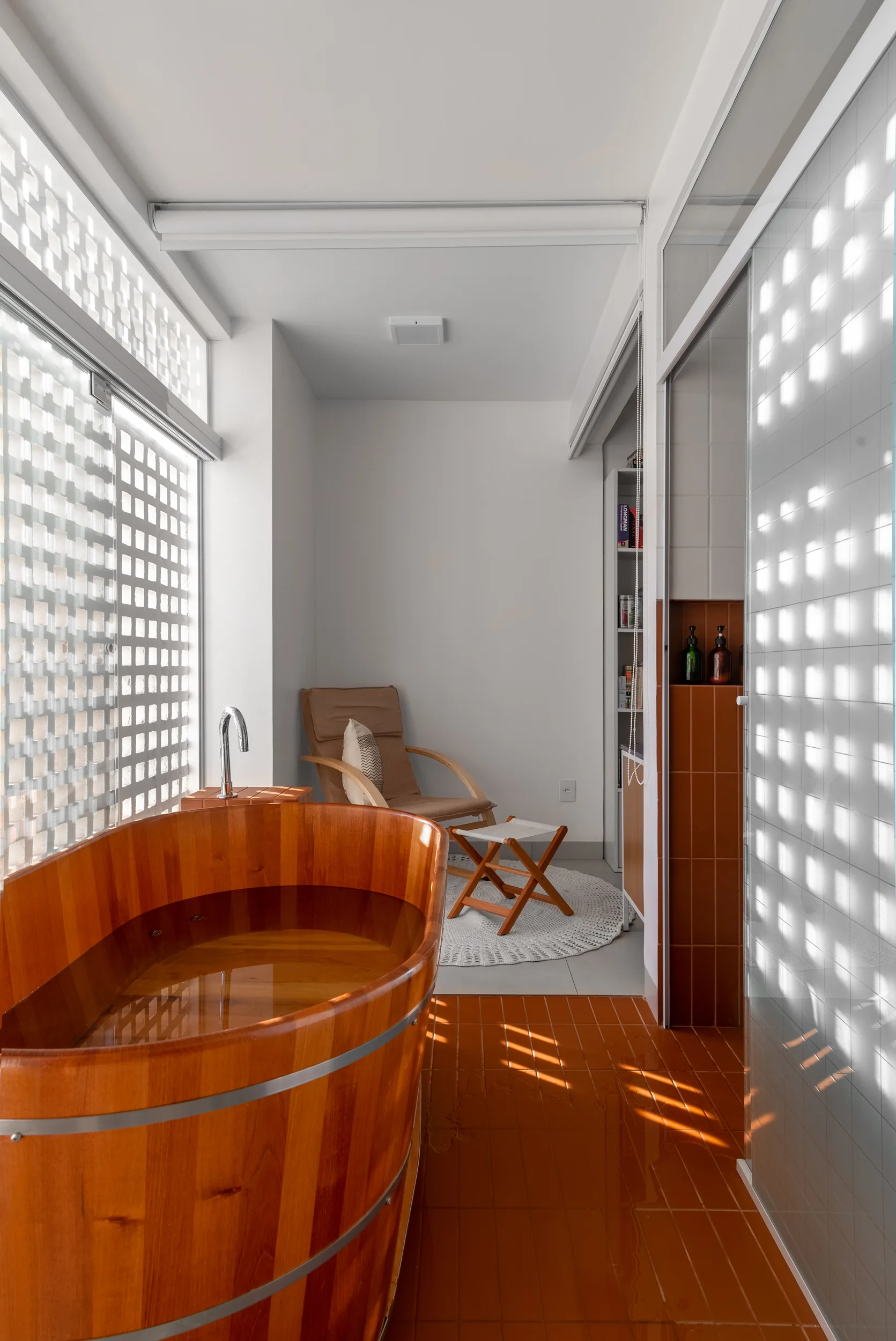 A bright bathroom features a wooden oval soaking tub on terracotta floor tiles next to a window with a white grid privacy screen.