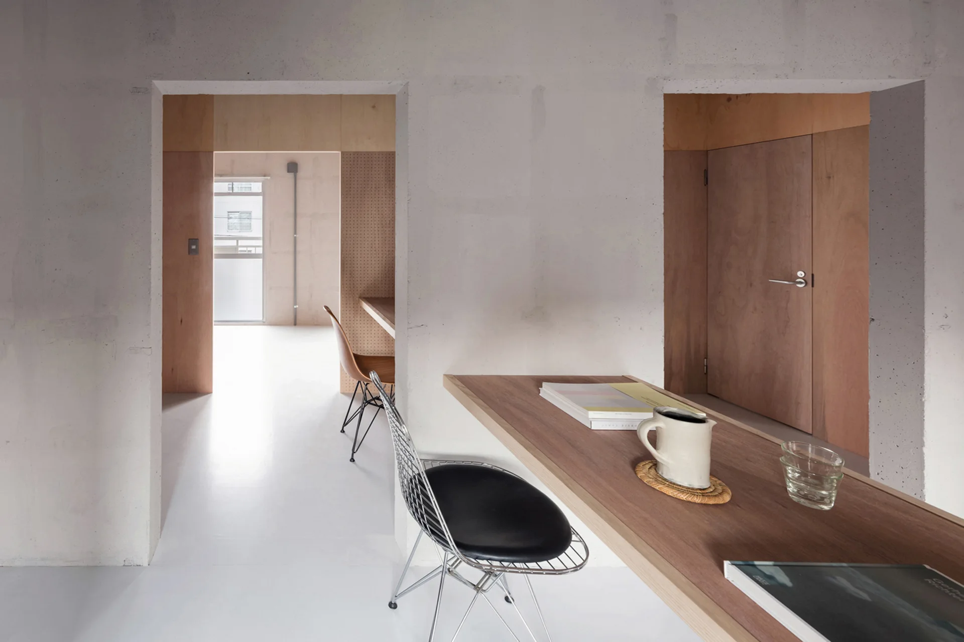 A minimalist office space features a built-in wooden desk, raw concrete walls, open doorways, and a wire mesh chair with a black cushion.