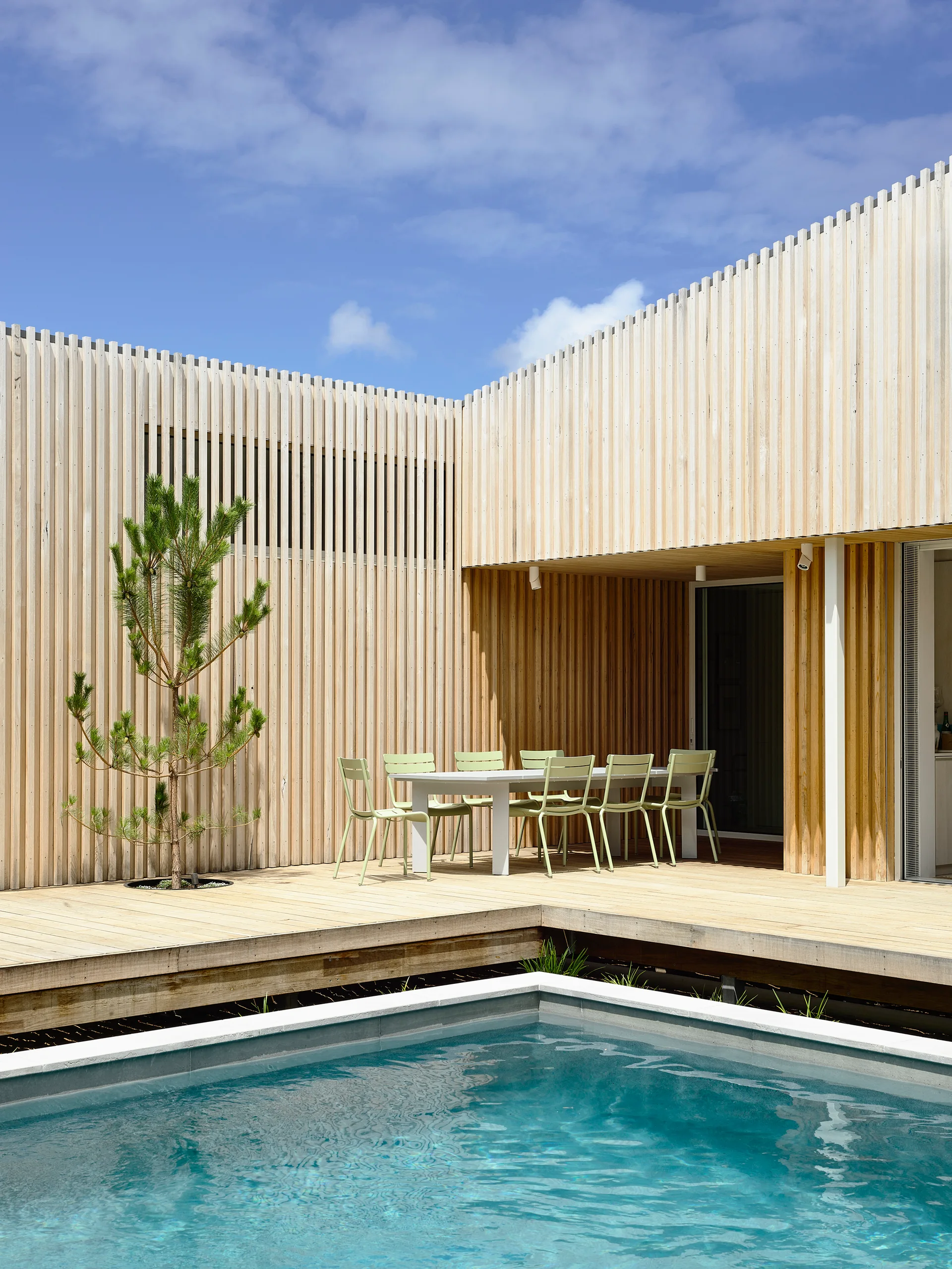 A modern outdoor patio featuring a wooden deck with a swimming pool and a dining set surrounded by vertical slatted wood walls.