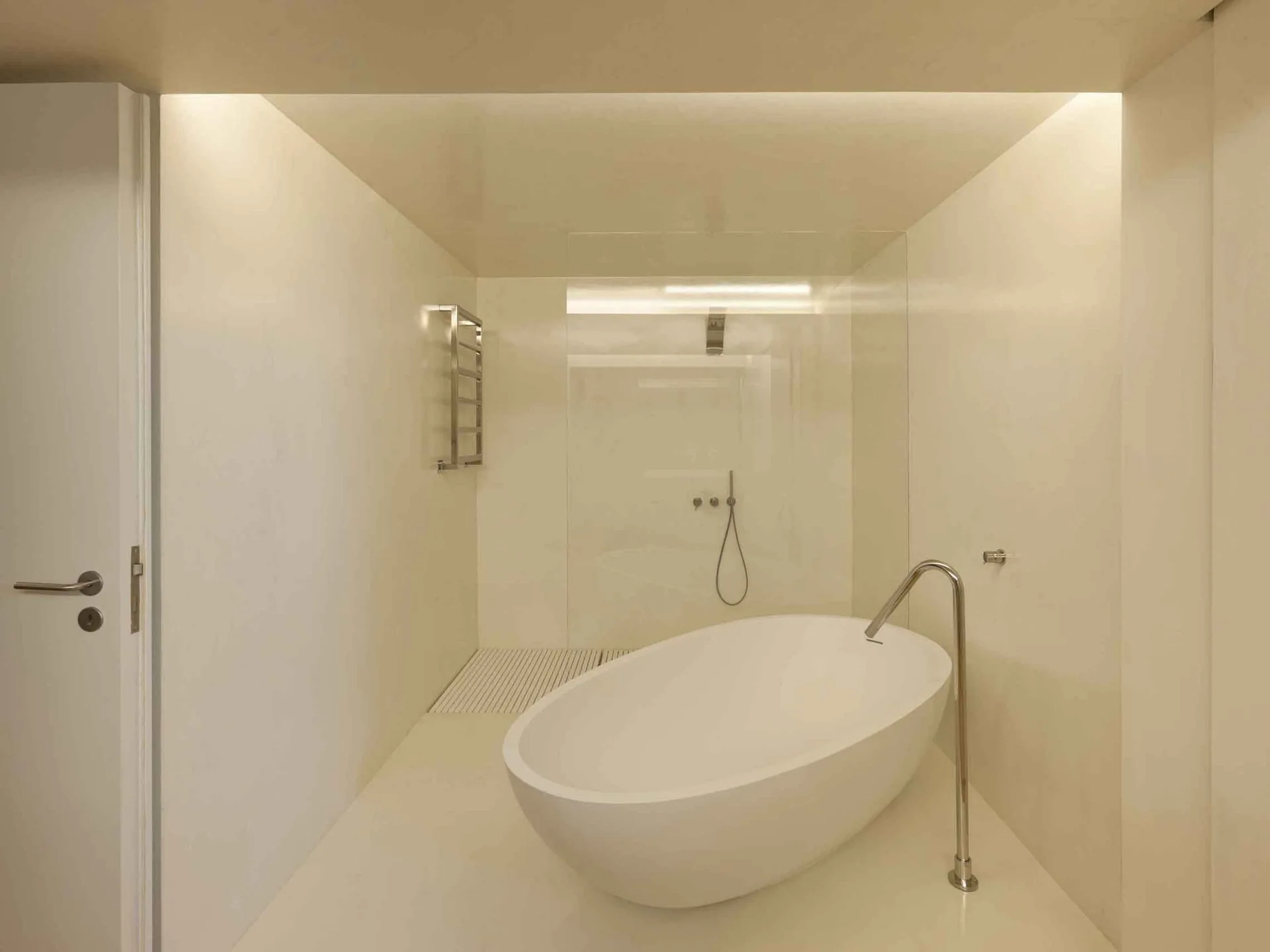 A minimalist all-white bathroom features a curved freestanding bathtub with a floor-mounted chrome faucet next to a glass-partitioned shower area.