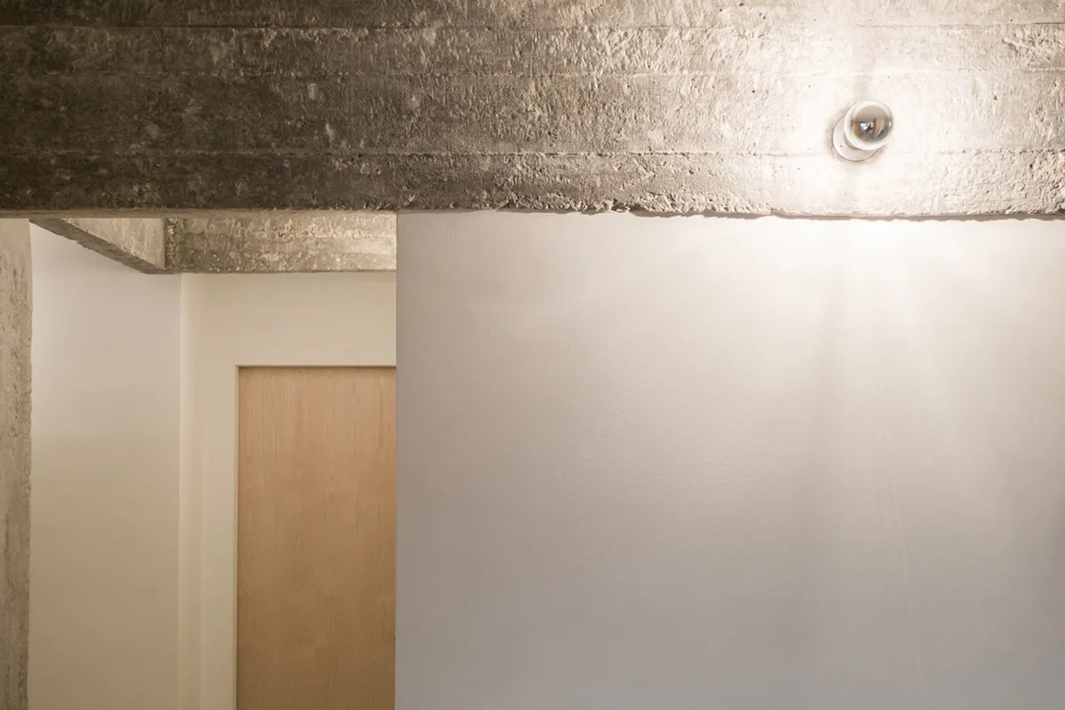 A minimalist hallway features a rugged exposed concrete ceiling beam with a mounted spotlight illuminating smooth white walls.