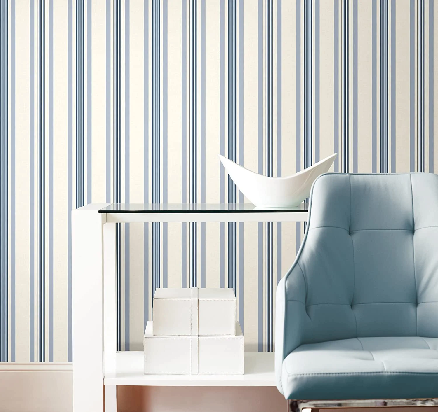 A light blue tufted armchair and a white glass-top console table stand in front of a wall with blue and cream vertical stripes.