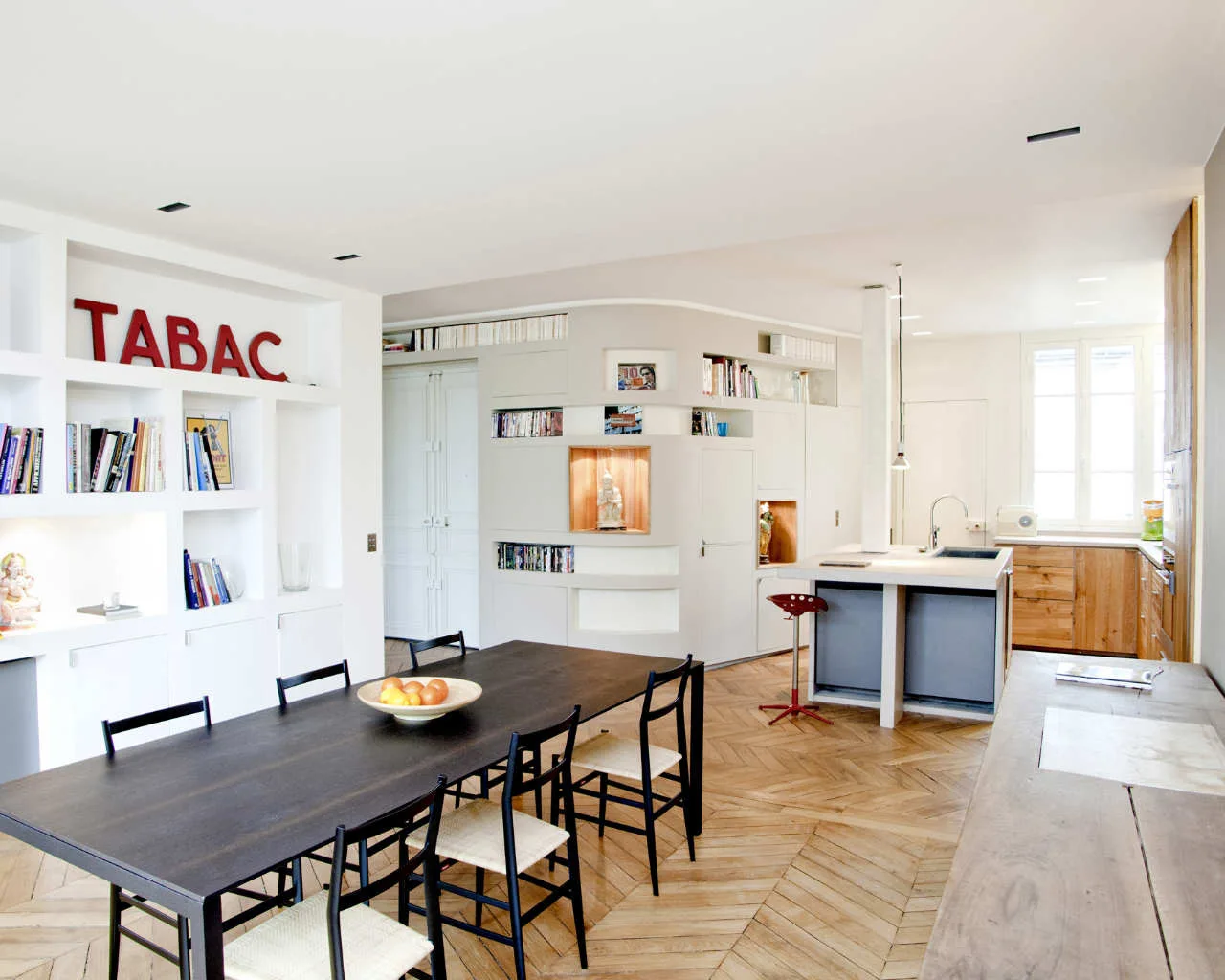 An open-plan dining area features a dark wood table, black chairs, and a white built-in bookcase wall on light herringbone wood floors.