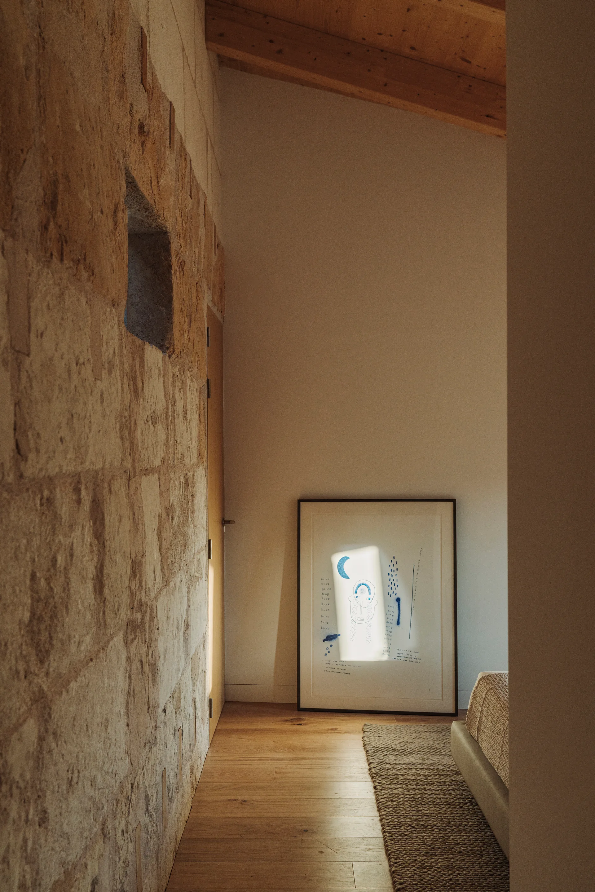 A bedroom hallway features a rough stone wall on one side, wood floors, and a framed blue graphic art piece leaning against a plain cream wall.