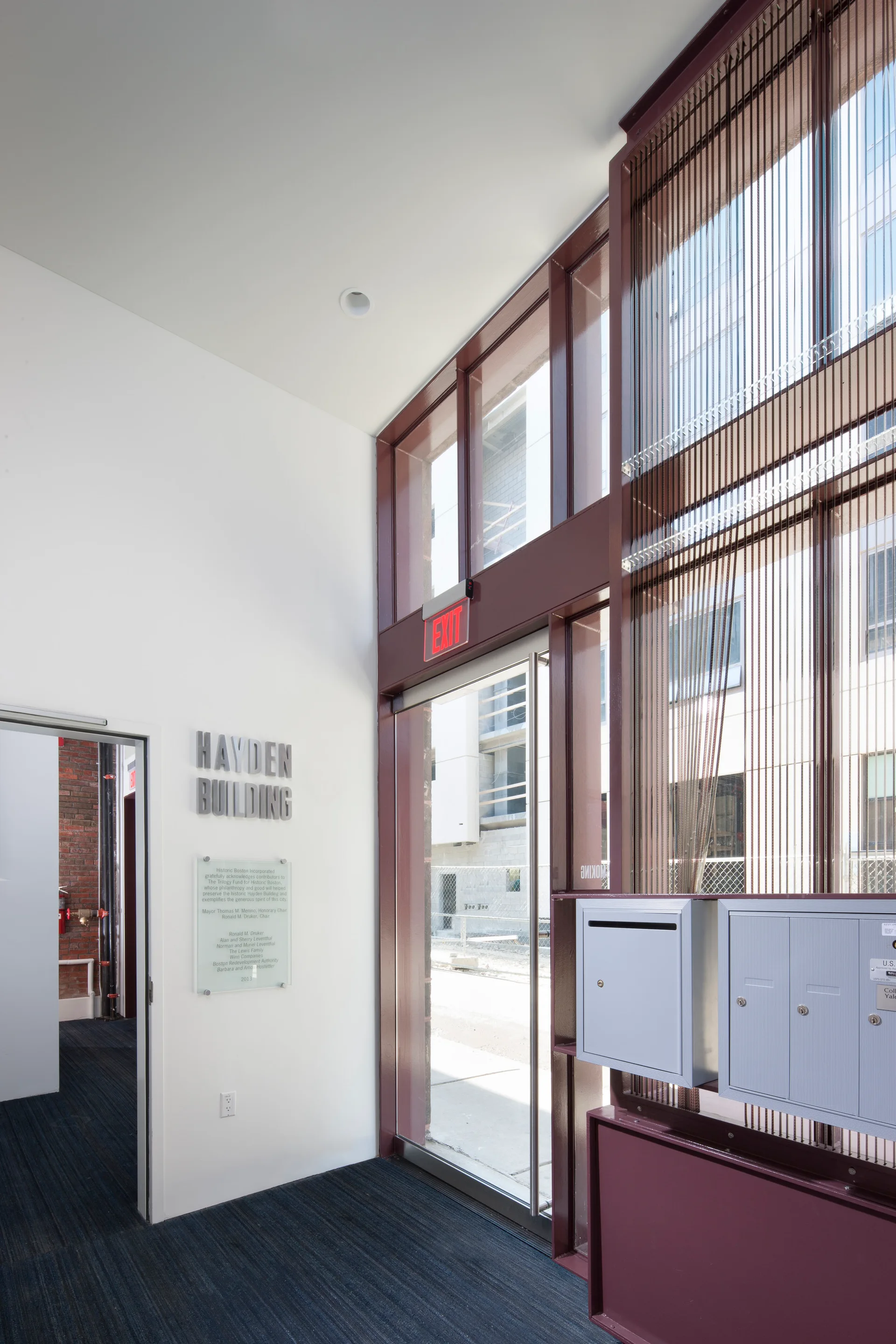 A modern lobby entryway features a large glass door with maroon metal framing, grey wall-mounted mailboxes, and a white wall with building signage.
