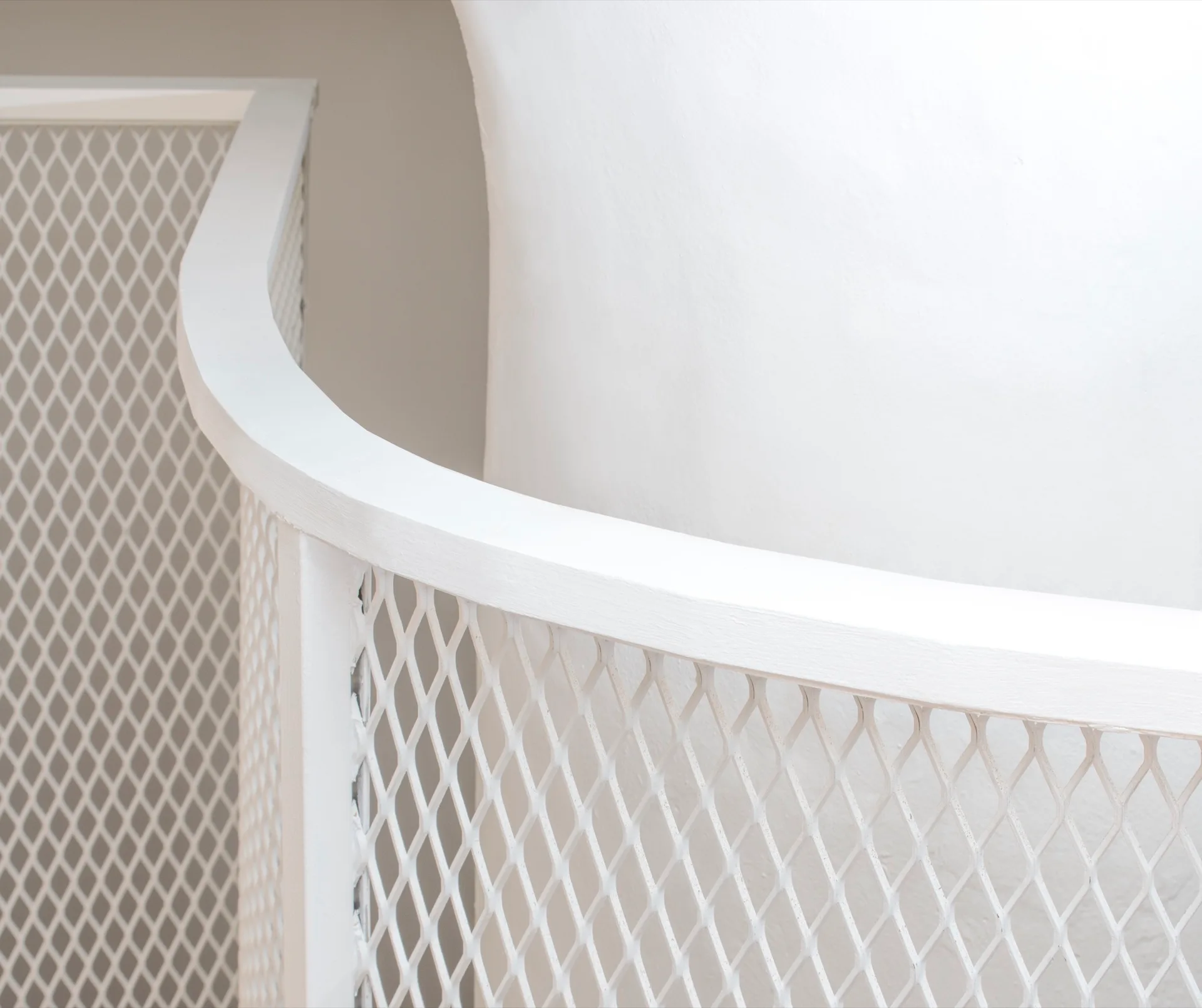 A close-up view of a curved white metal staircase railing featuring a diamond mesh pattern against a smooth white wall.