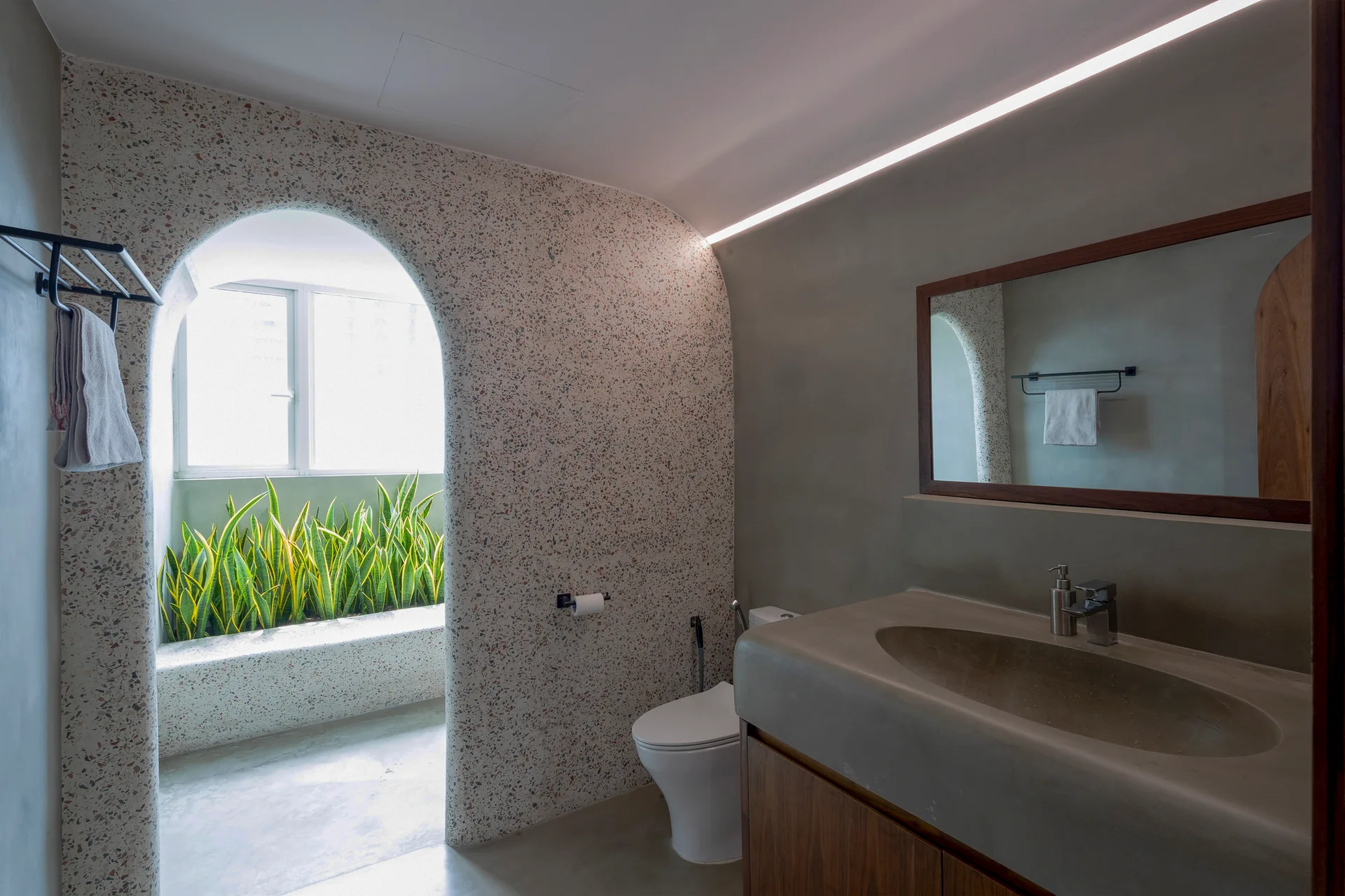 A modern bathroom features a speckled terrazzo archway leading to a plant-filled alcove, alongside a concrete sink vanity and wood-framed mirror.