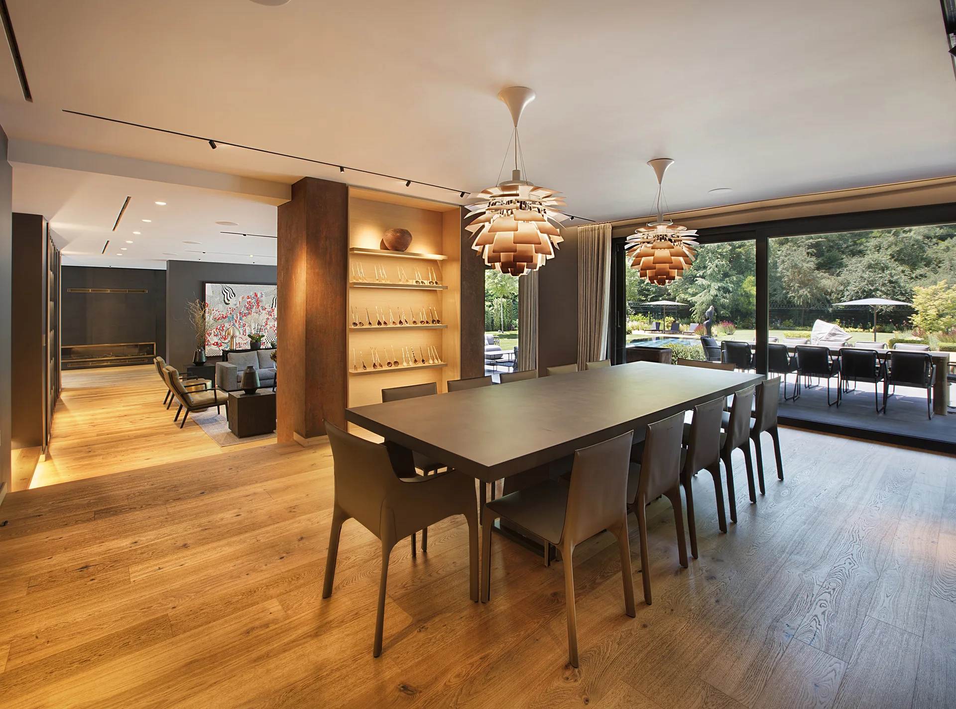 A spacious dining room featuring a long dark table with chairs, two tiered sculptural pendant lights, lighted display shelving, and wide-plank wood flooring.