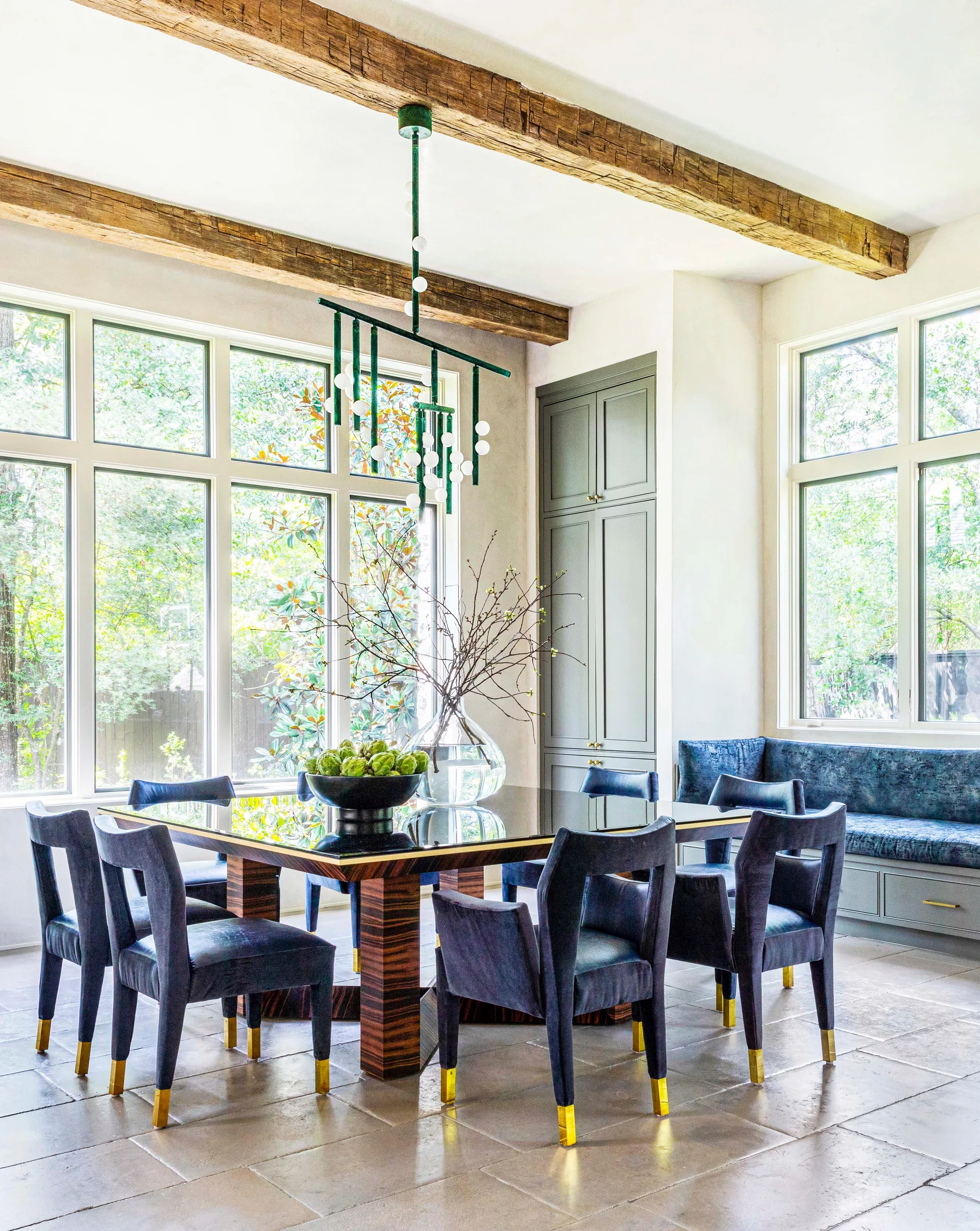 Navy upholstered chairs surround a dark glass-top table beneath exposed wood ceiling beams and a green modern chandelier in a bright dining room.
