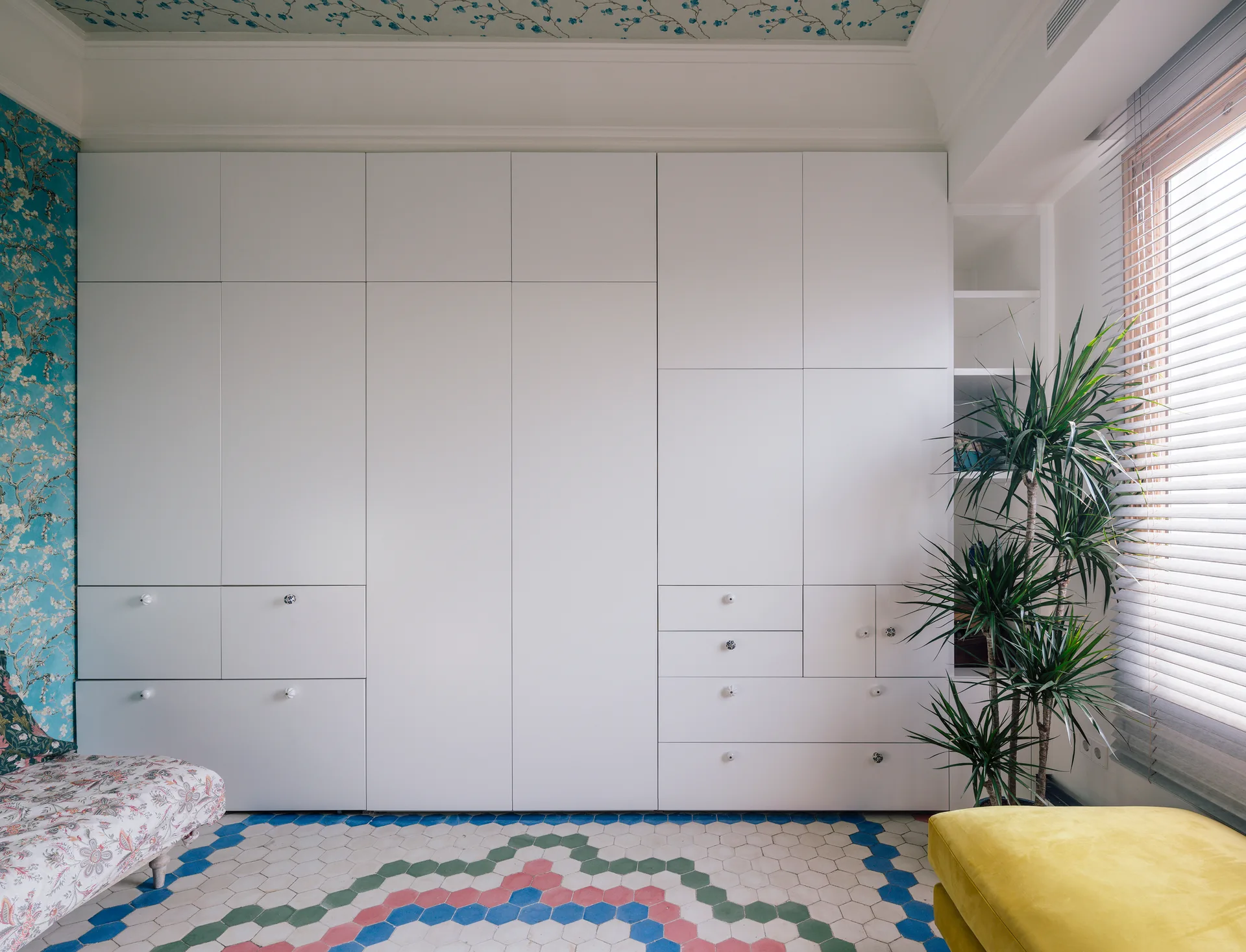 A white built-in wardrobe stands next to blue floral wallpaper, featuring multicolored hexagonal floor tiles and a potted plant by the window.