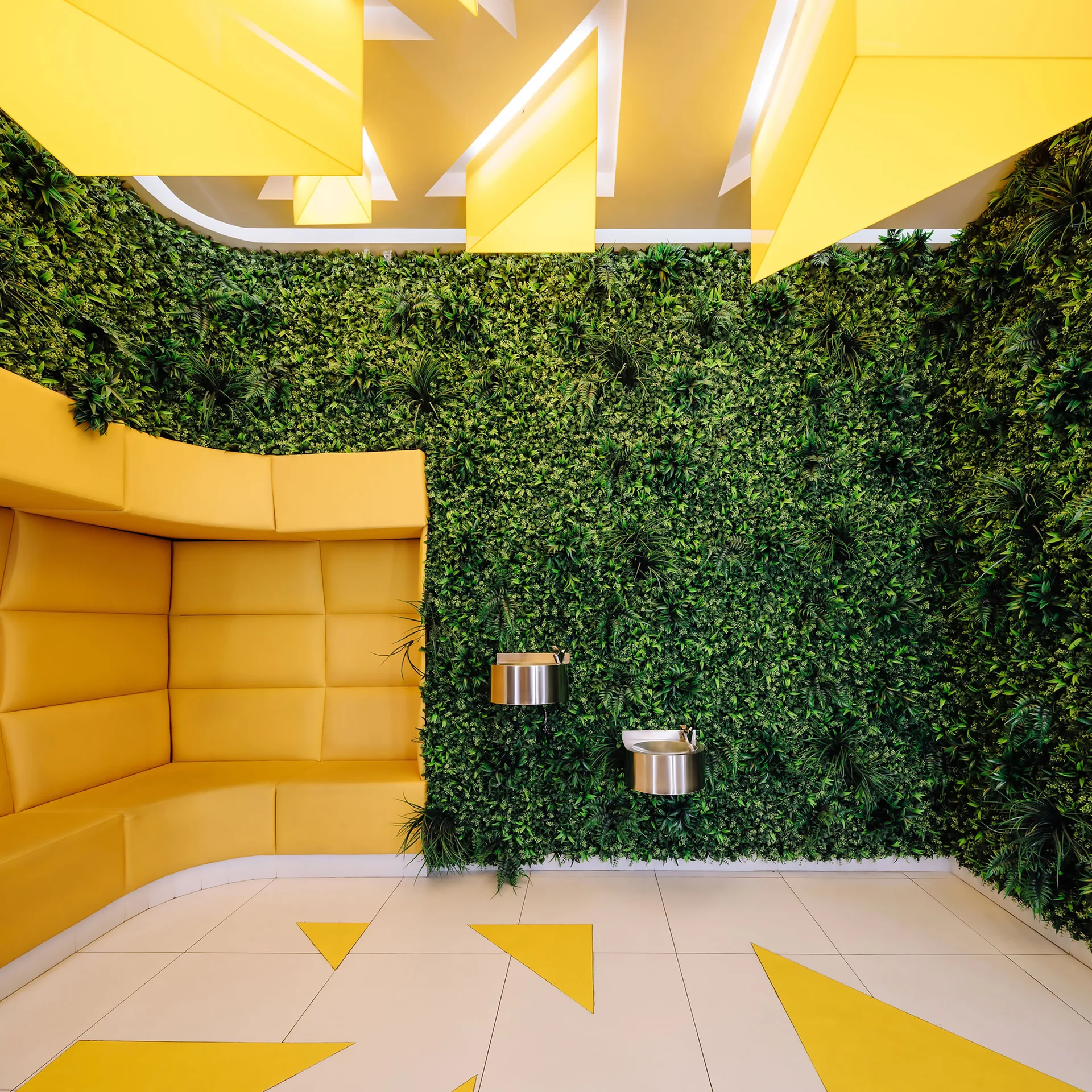 A commercial indoor lounge features a dense green wall of foliage, a bright yellow curved modular sofa, and geometric yellow ceiling panels.