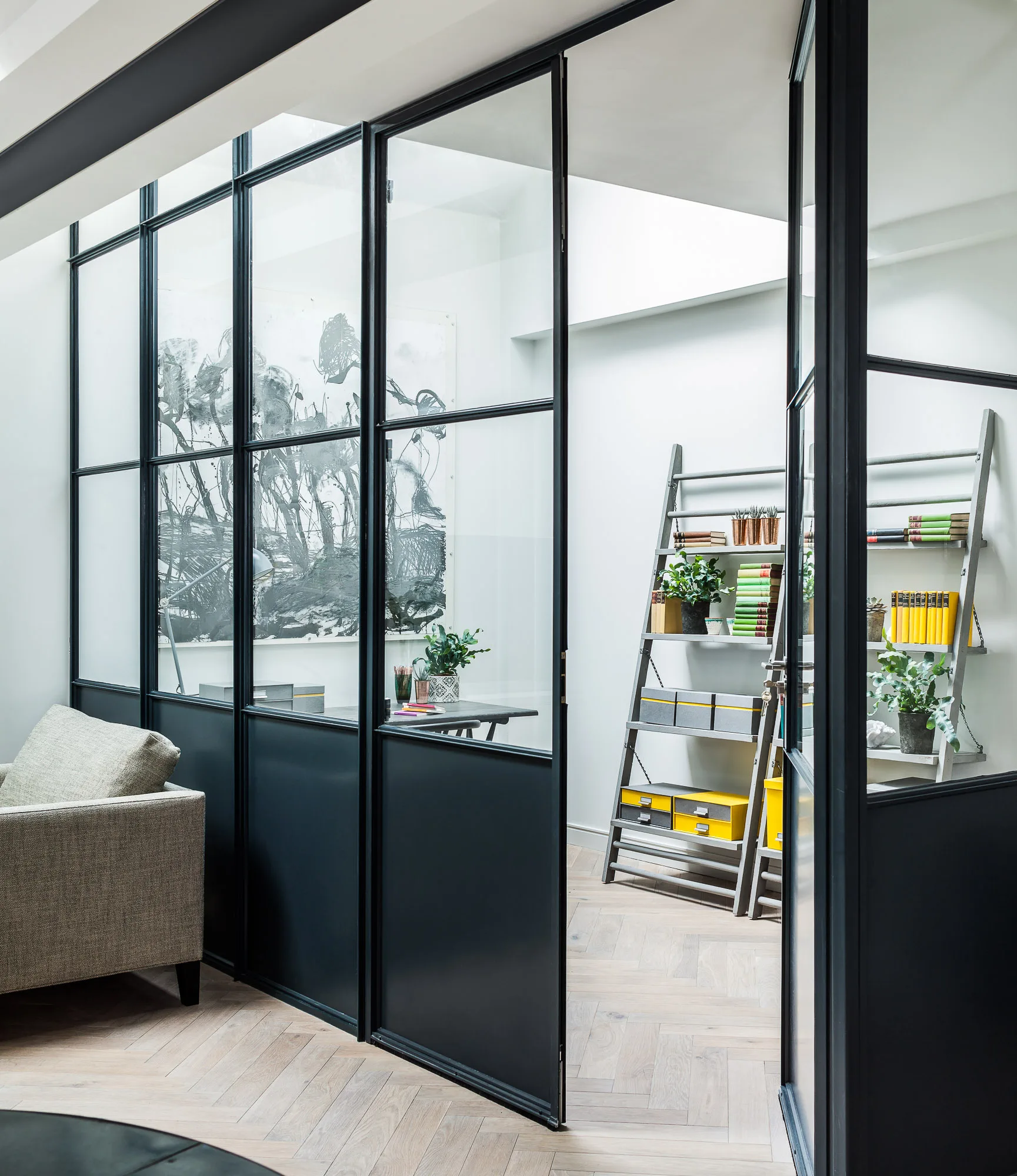 A black steel-framed glass partition separates a living space from a home office featuring ladder shelving and herringbone parquet wood floors.