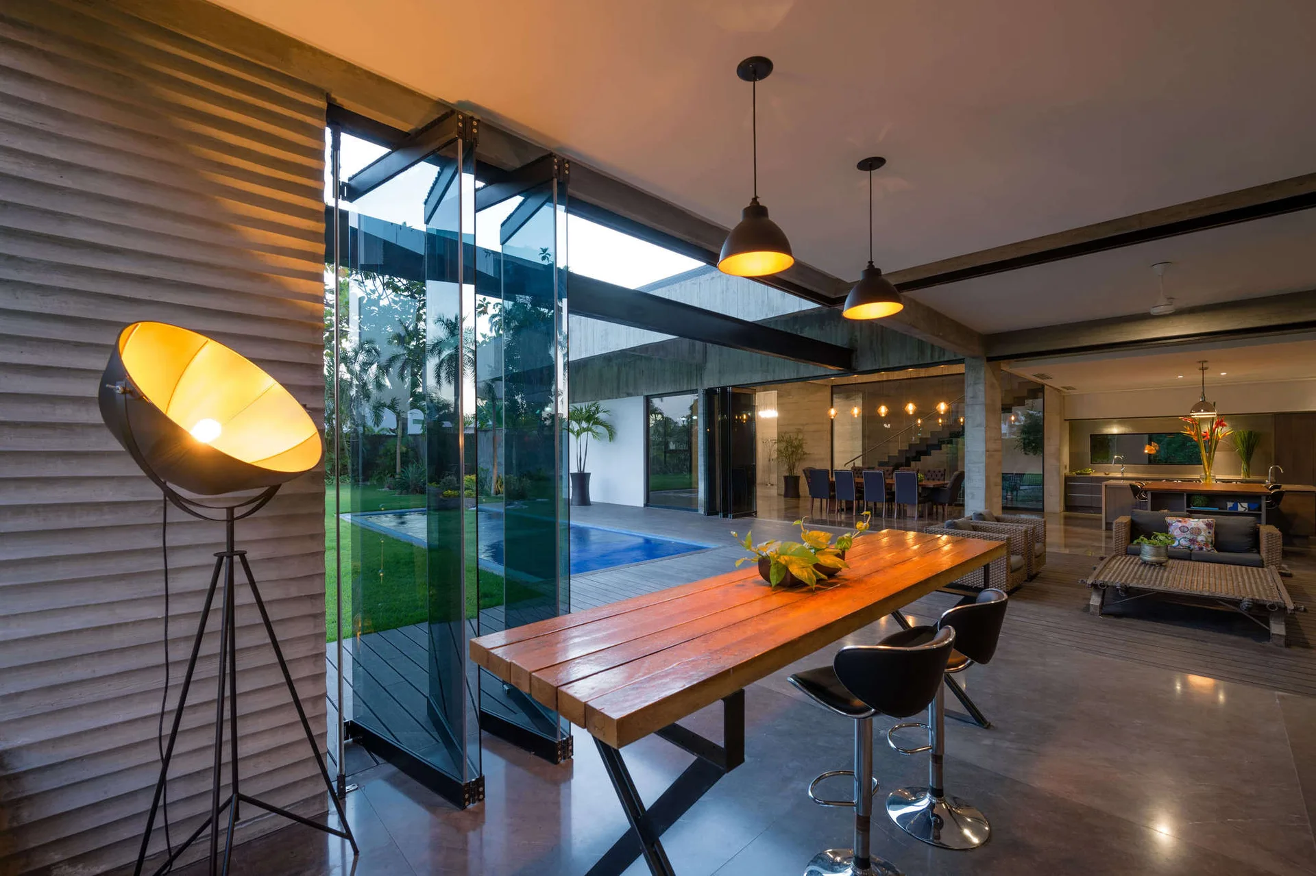 A modern dining space featuring a long wooden table, black bar stools, a ribbed concrete wall, and folding glass doors that open to an outdoor pool.