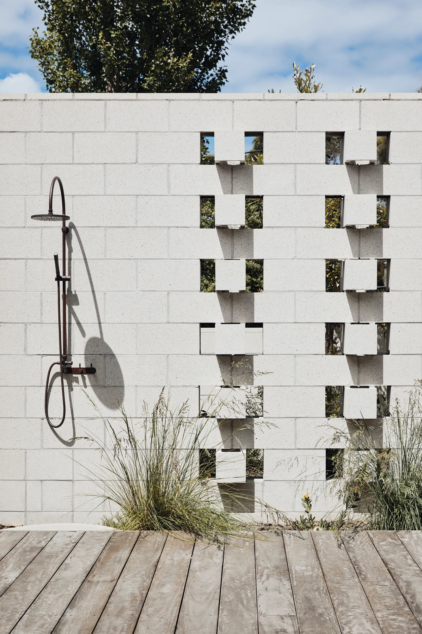 A wall-mounted black metal outdoor shower set against a white cinder block wall with open geometric cutouts above a weathered wood deck.