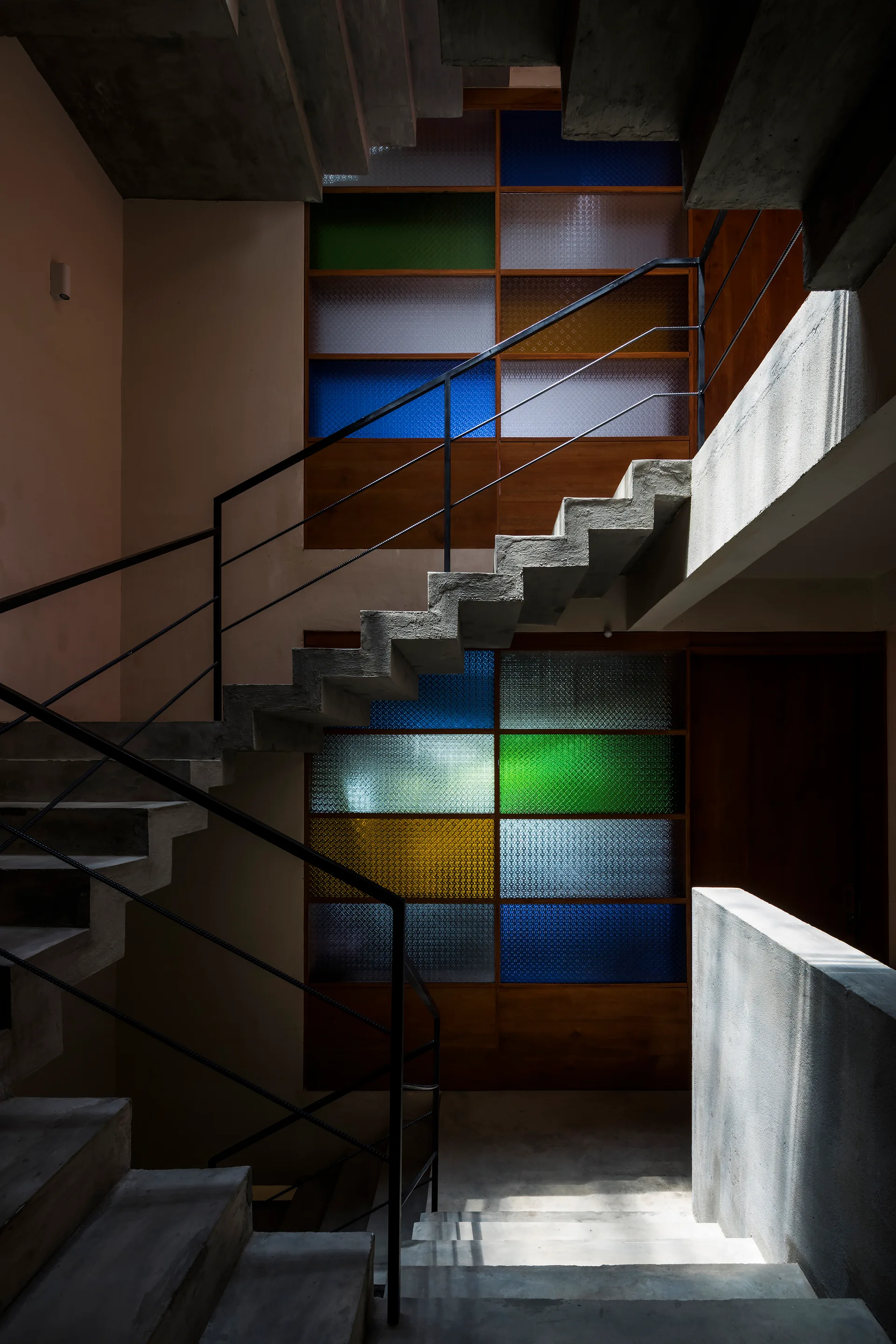 A raw concrete staircase wraps around a large wood-framed partition wall filled with yellow, blue, and green textured glass panels.